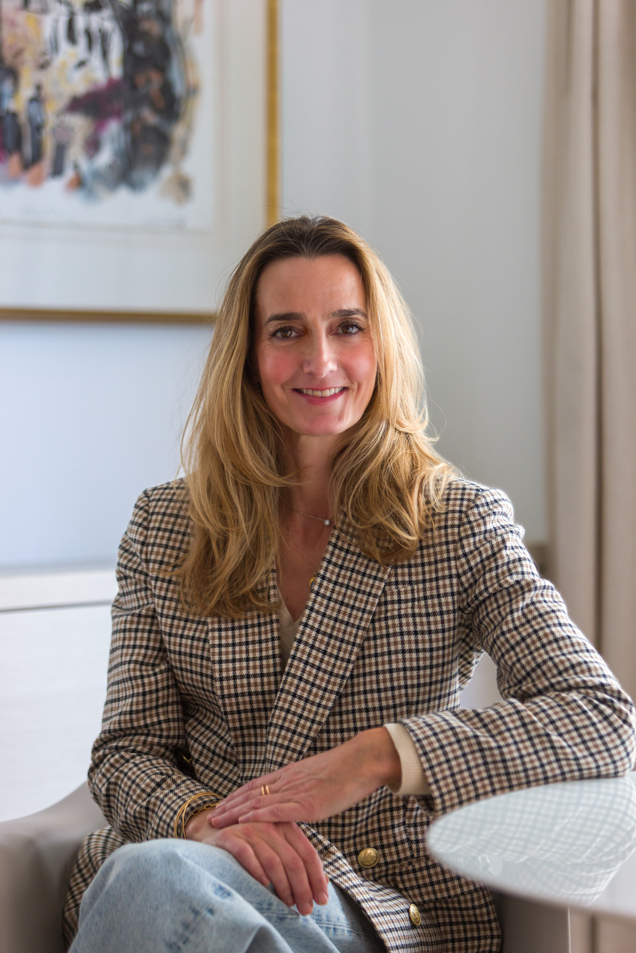 A woman with long blonde hair sitting at a white table, smiling, wearing a checkered blazer and light jeans, in a well-lit room with art on the wall.