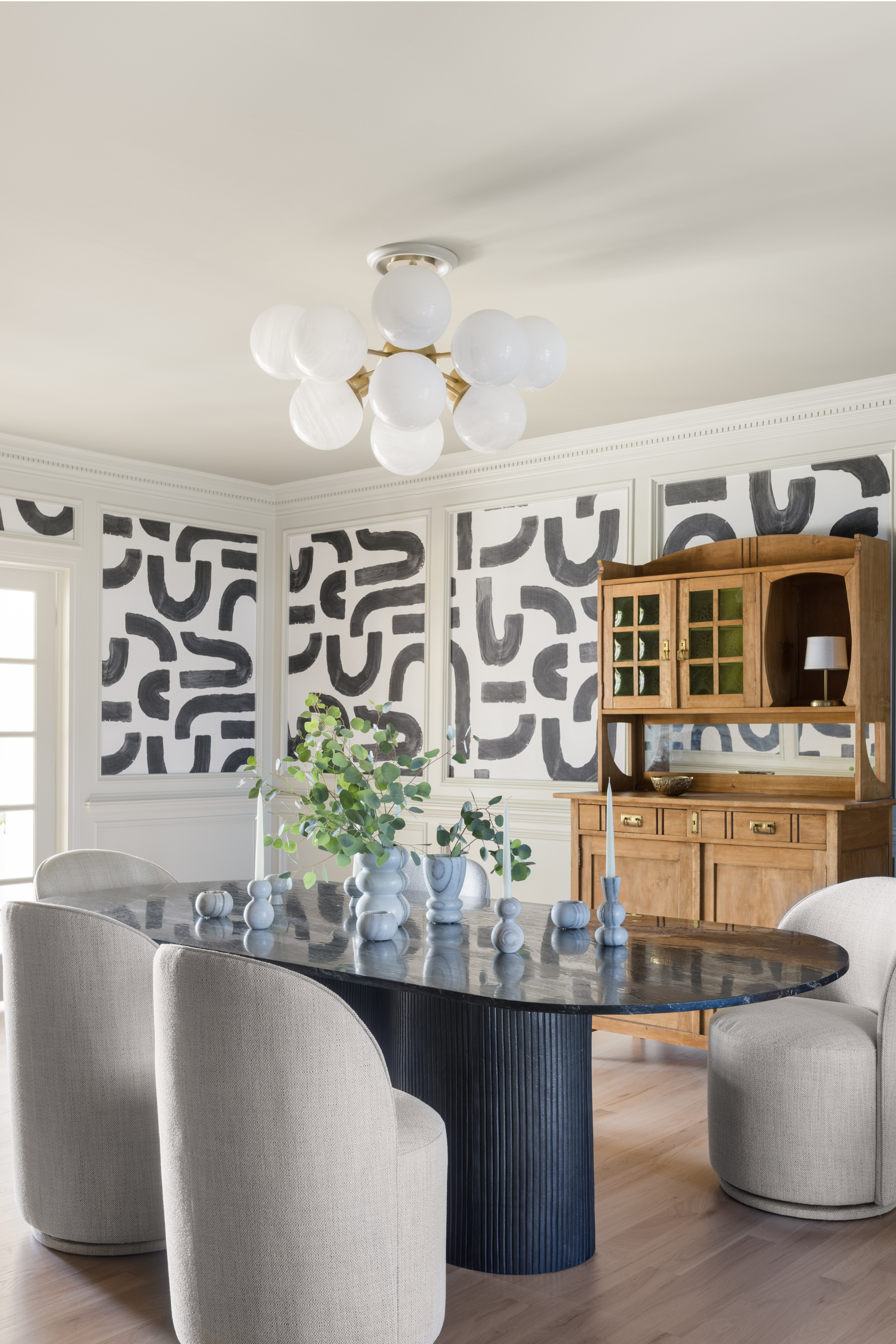 A dining room with four beige upholstered chairs around a dark, oval marble table with a vase of green leafy branches. The room has black and white abstract wall art, a wooden hutch, and a modern white ceiling light fixture.