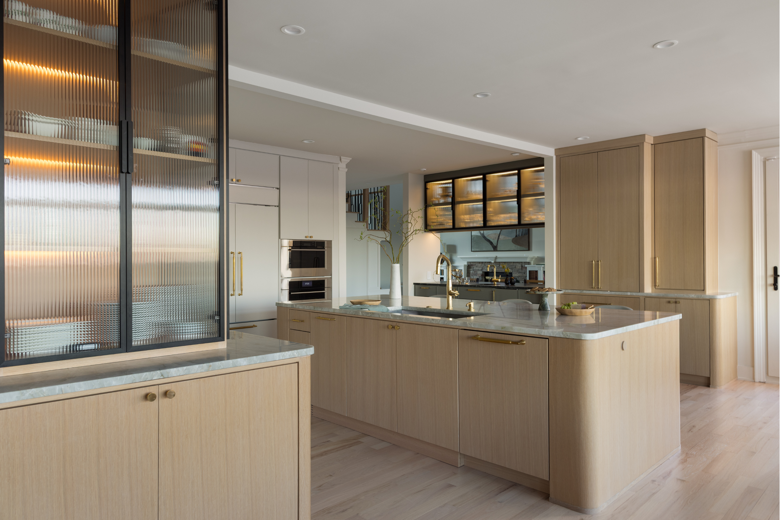 Modern kitchen with light wood cabinets, marble countertops, and gold hardware. Features a large kitchen island with a sink, a built-in oven, and display cabinets with lighting.