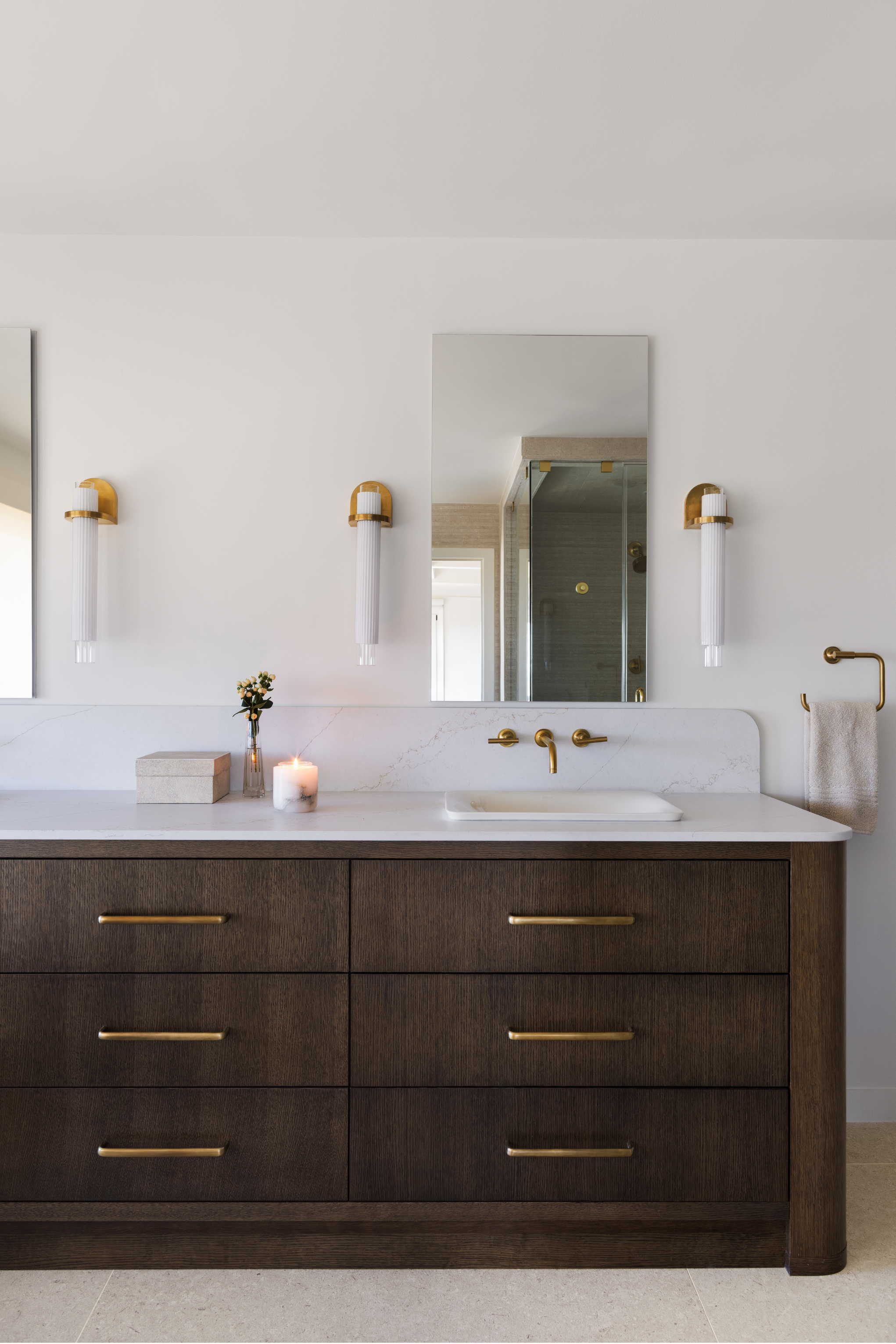 Bathroom vanity with white countertop, dark wood drawers with gold handles, wall-mounted gold faucet, large mirror, wall lights with gold accents, small flower vase, lit candle, towel hanging on gold towel rack.