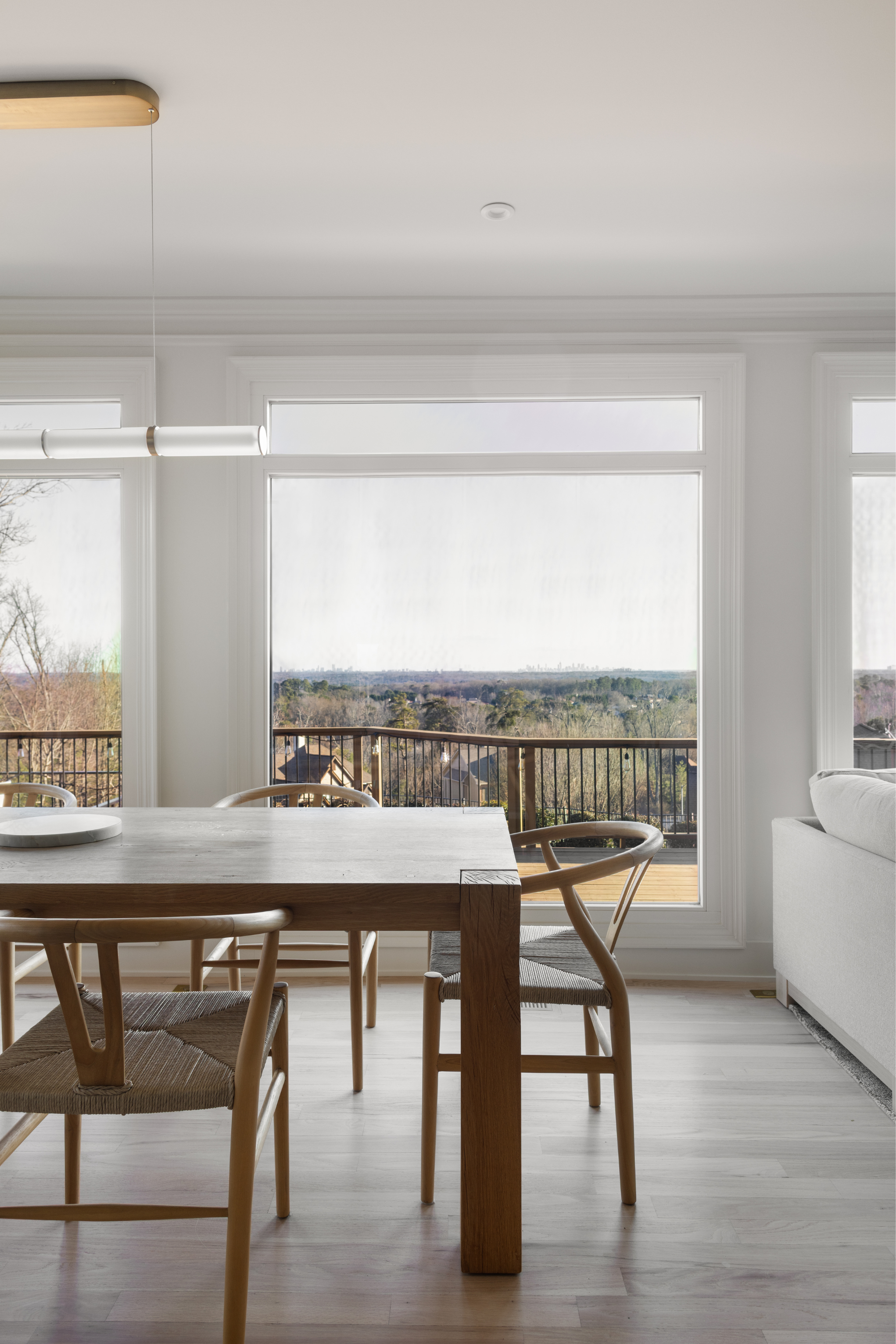 Bright dining area with wooden table and chairs, large windows showing a scenic view of trees and sky outside, and a white sofa on the right.