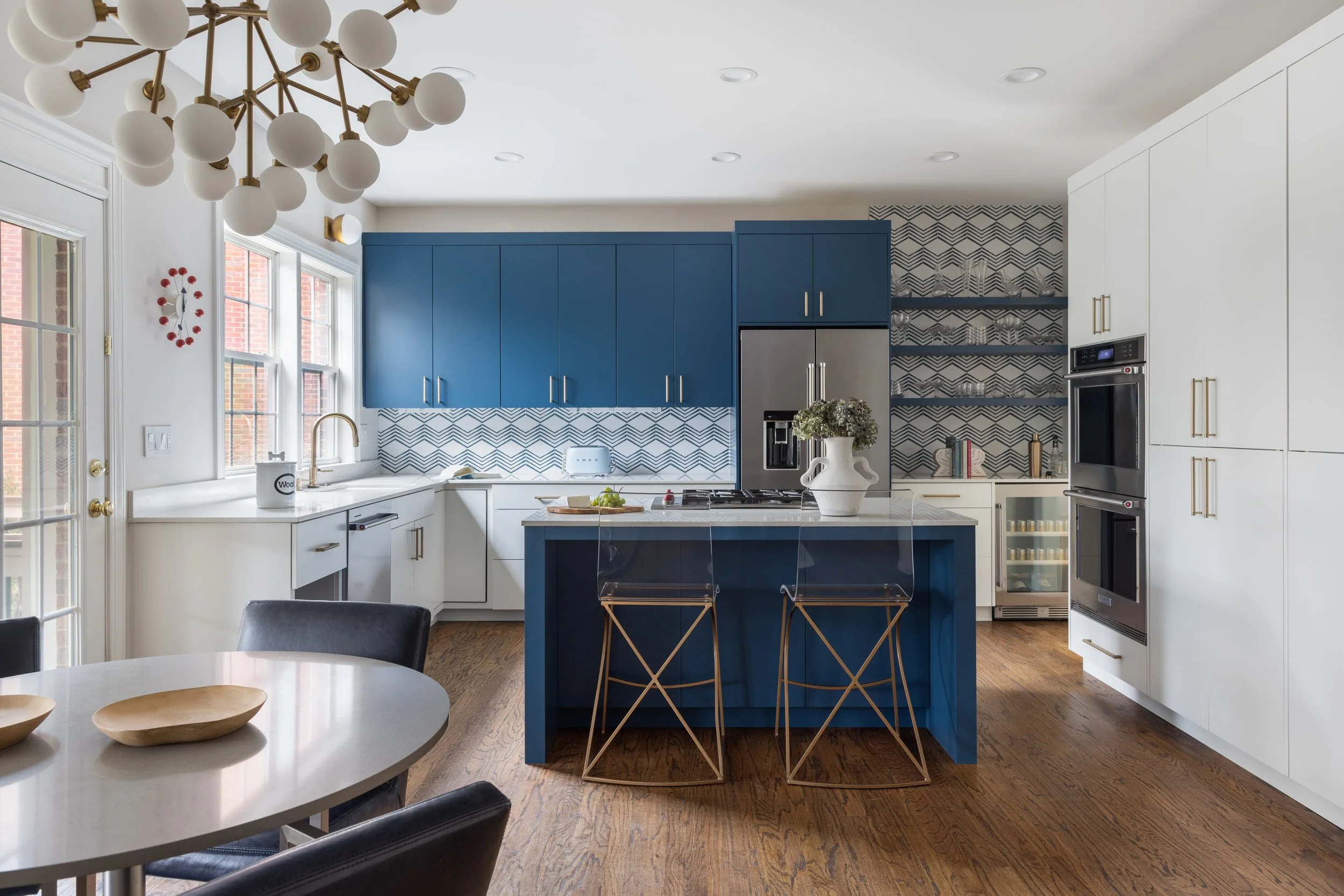 Kitchen with a blue and white color scheme, featuring blue upper cabinets, white lower cabinets, a blue kitchen island with two gold bar stools, a patterned backsplash, and a dining table with black chairs.