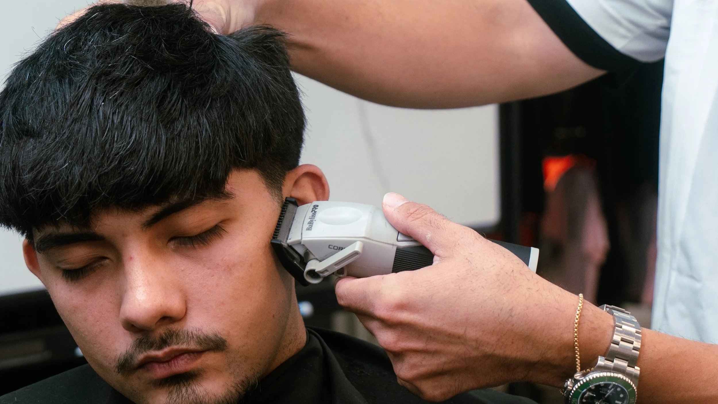 A young man with black hair receiving a haircut or grooming, with a barber using electric clippers on his hair. The young man has closed eyes, and a barber's hand is visible wearing a silver bracelet and wristwatch.