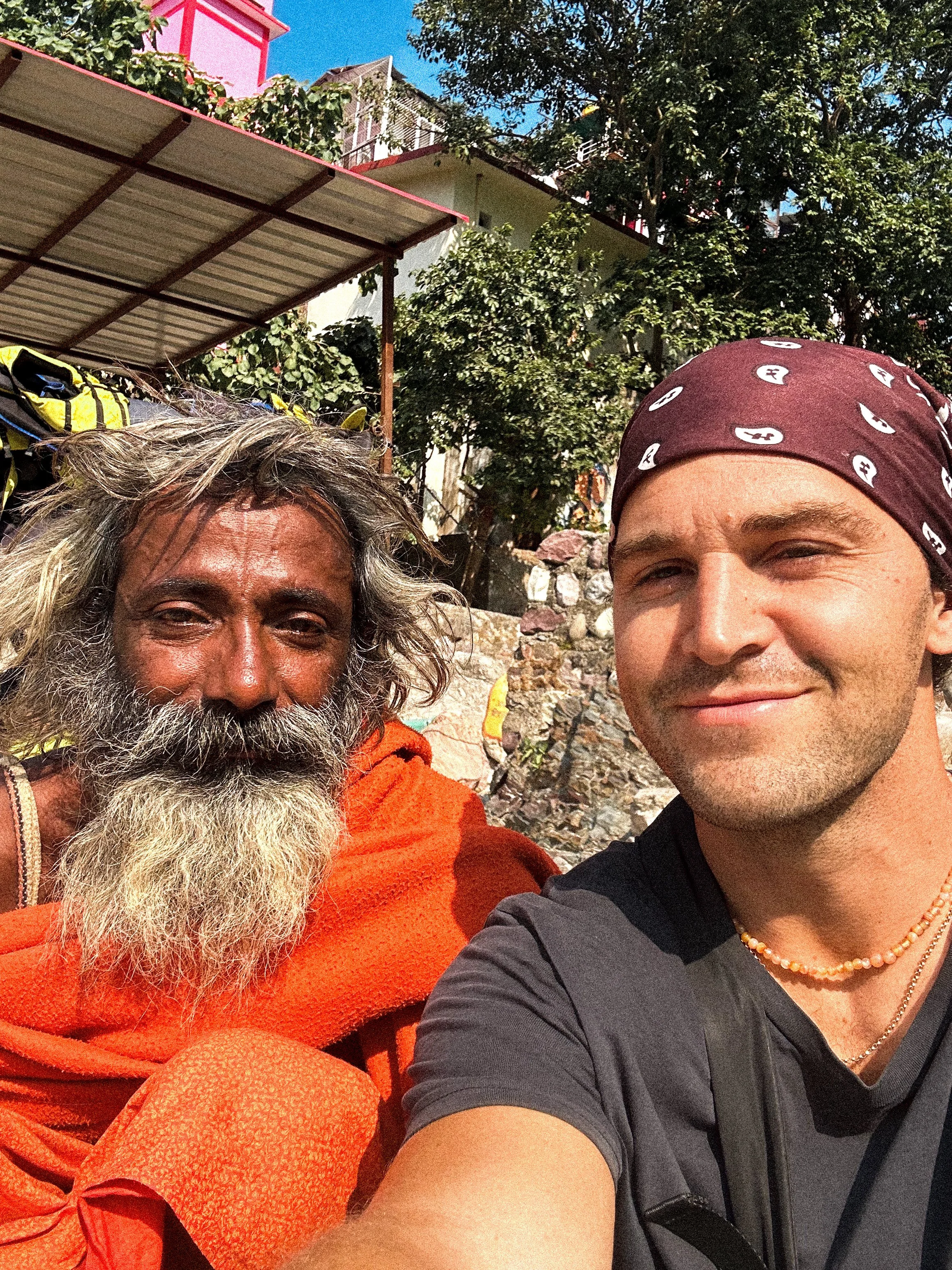 A selfie of two men outdoors: an elderly man with a long gray beard, wearing an orange robe, and a young man with a bandana, smiling. Background includes trees, a building, and a rusty roof structure.