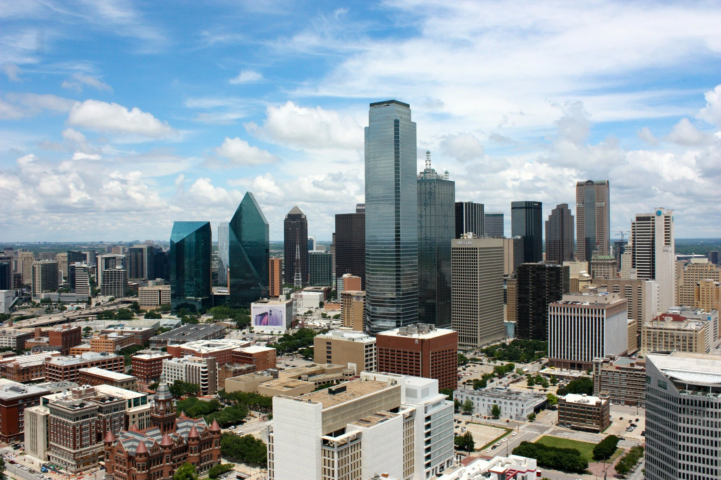 Aerial view of a downtown Dallas skyline with tall skyscrapers, partly cloudy sky, and streets below.