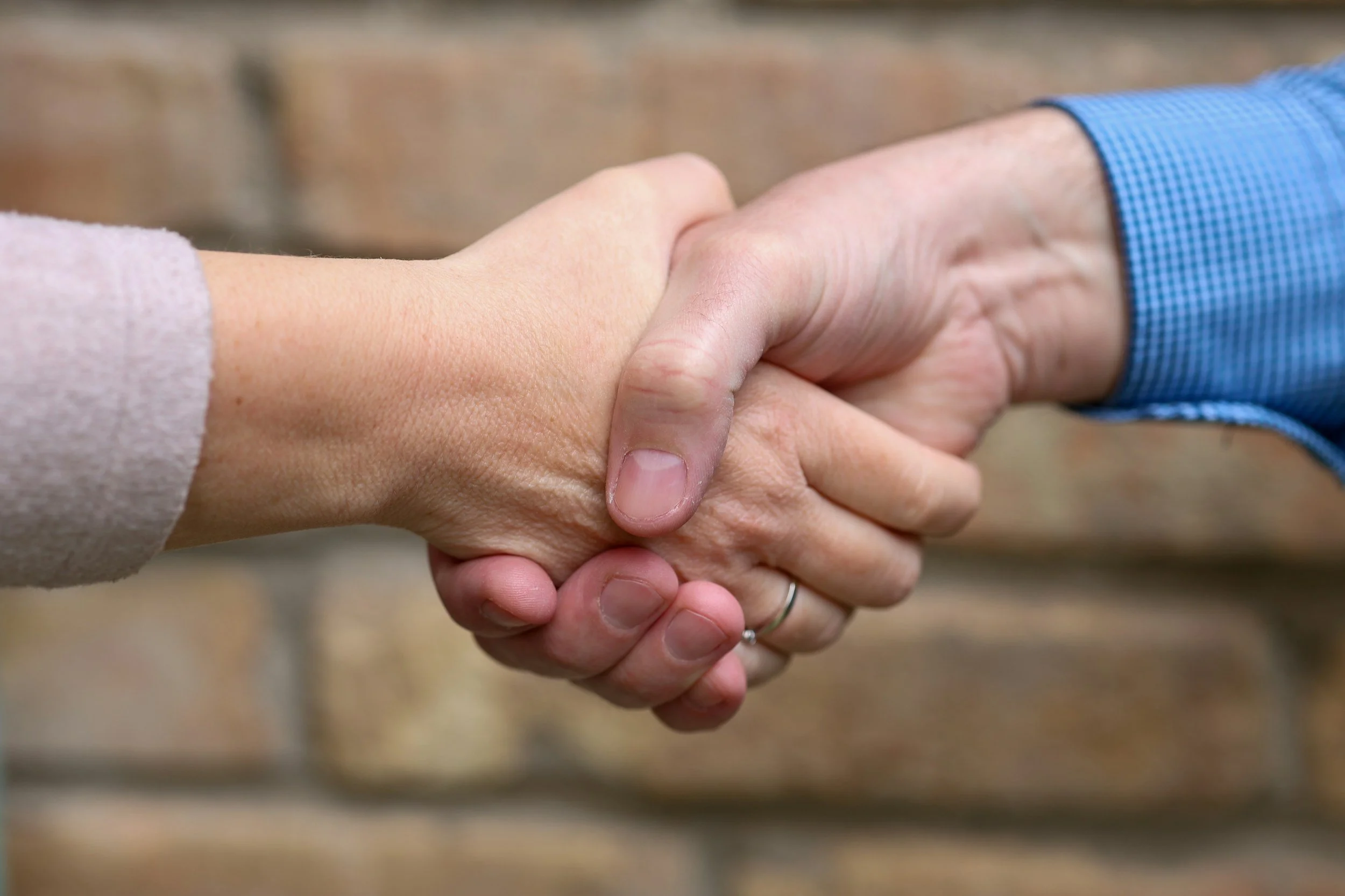 Close-up of two people shaking hands, one with a pink sweater and the other with a blue shirt, in front of a brick wall.