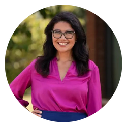 A woman with dark hair and glasses smiling outdoors, wearing a bright pink blouse.