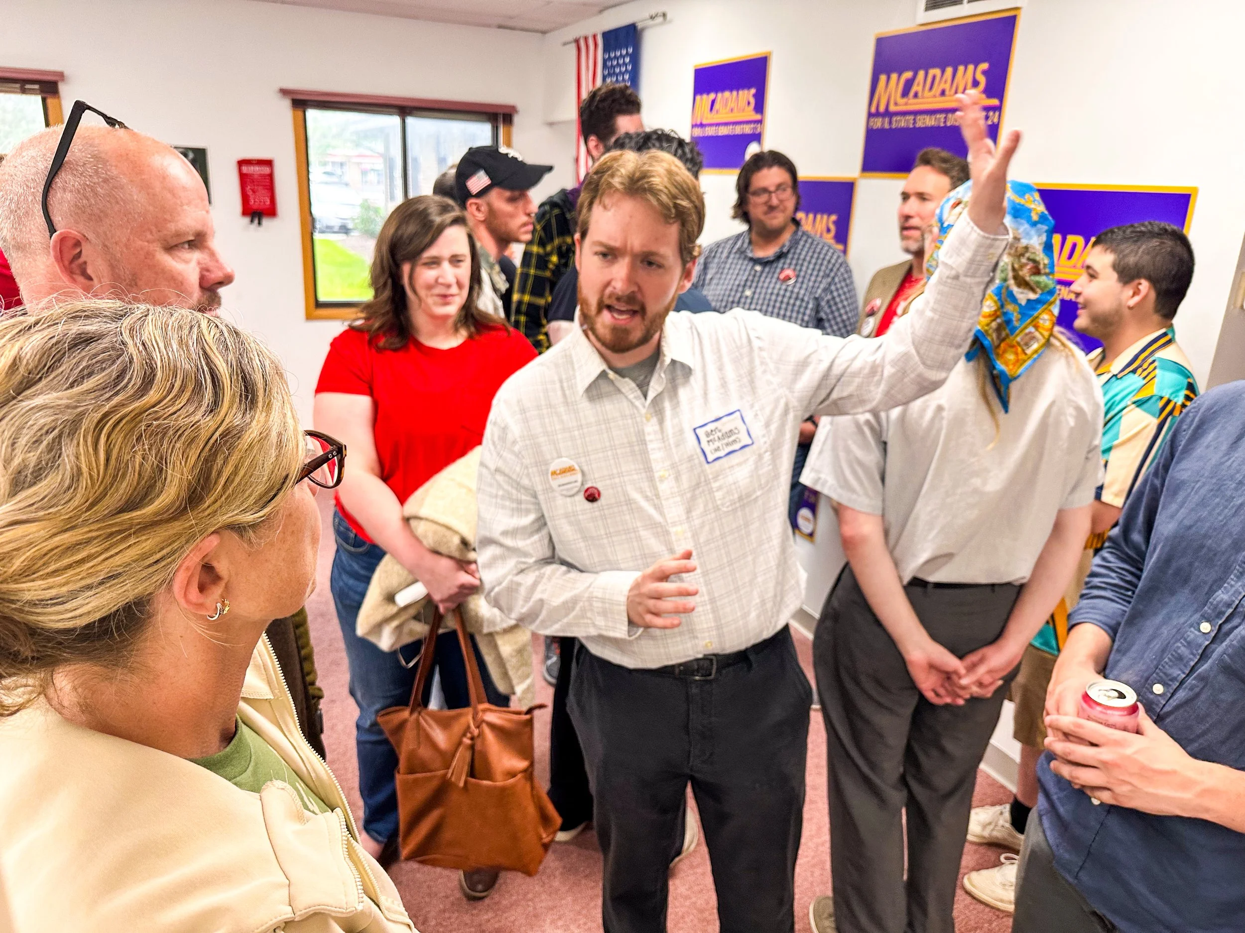 Group of people engaged in conversation at an indoor event, with a man gesturing with his hand while others listen. There are posters and windows in the background, and some attendees are holding drinks.