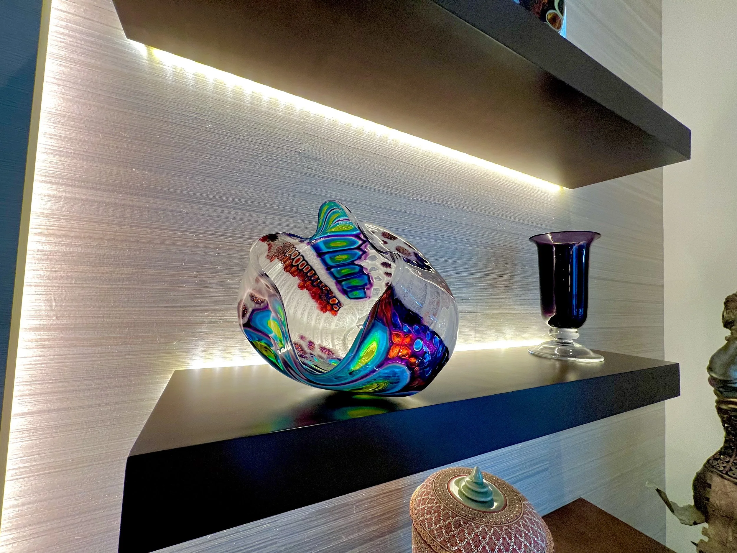Decorative glass art pieces on a black shelf, including a multi-colored swirling glass orb and a dark purple glass vase, with a textured wall background and soft lighting.