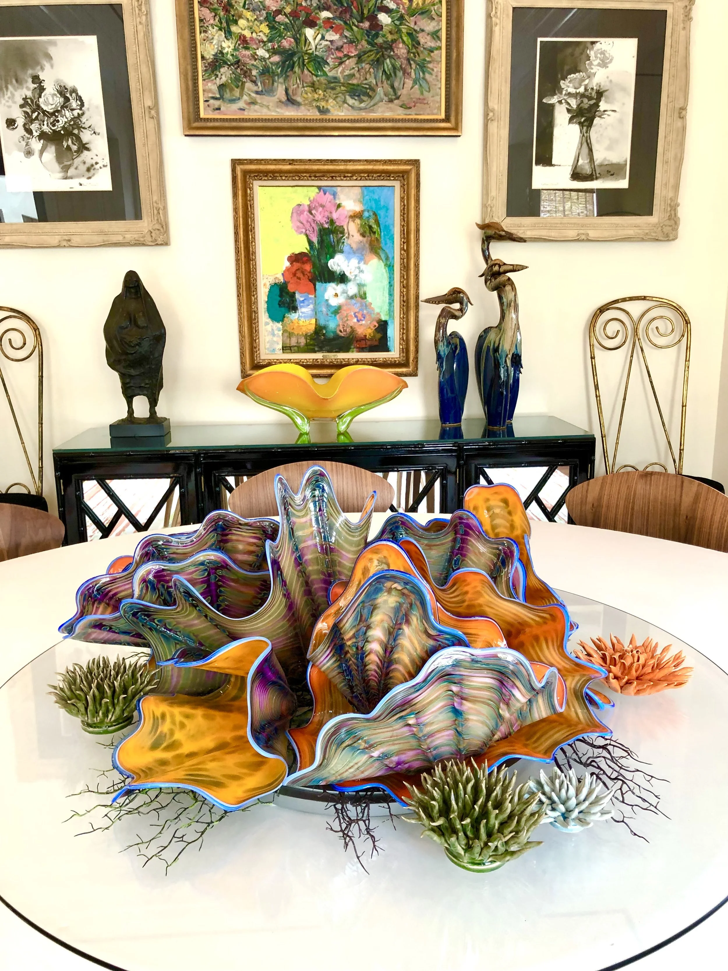 Colorful glass sculpture centerpiece on a white table, surrounded by decorative plants. Background features framed artwork and ceramics, including a multi-colored abstract painting, black and white floral prints, and sculptures of birds and a woman.