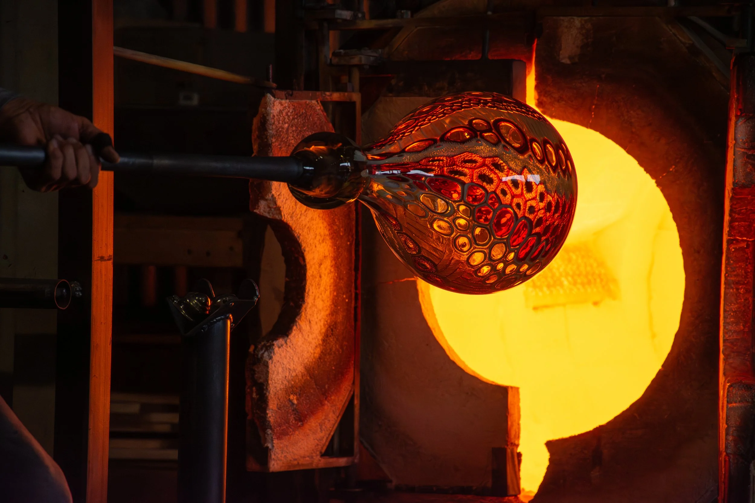 A person holding a glassblowing pipe with a molten glass piece in a glassblowing studio. The molten glass is glowing orange and red, shaped like a decorative sphere with intricate patterns, in front of a furnace with a bright yellow-orange flame.