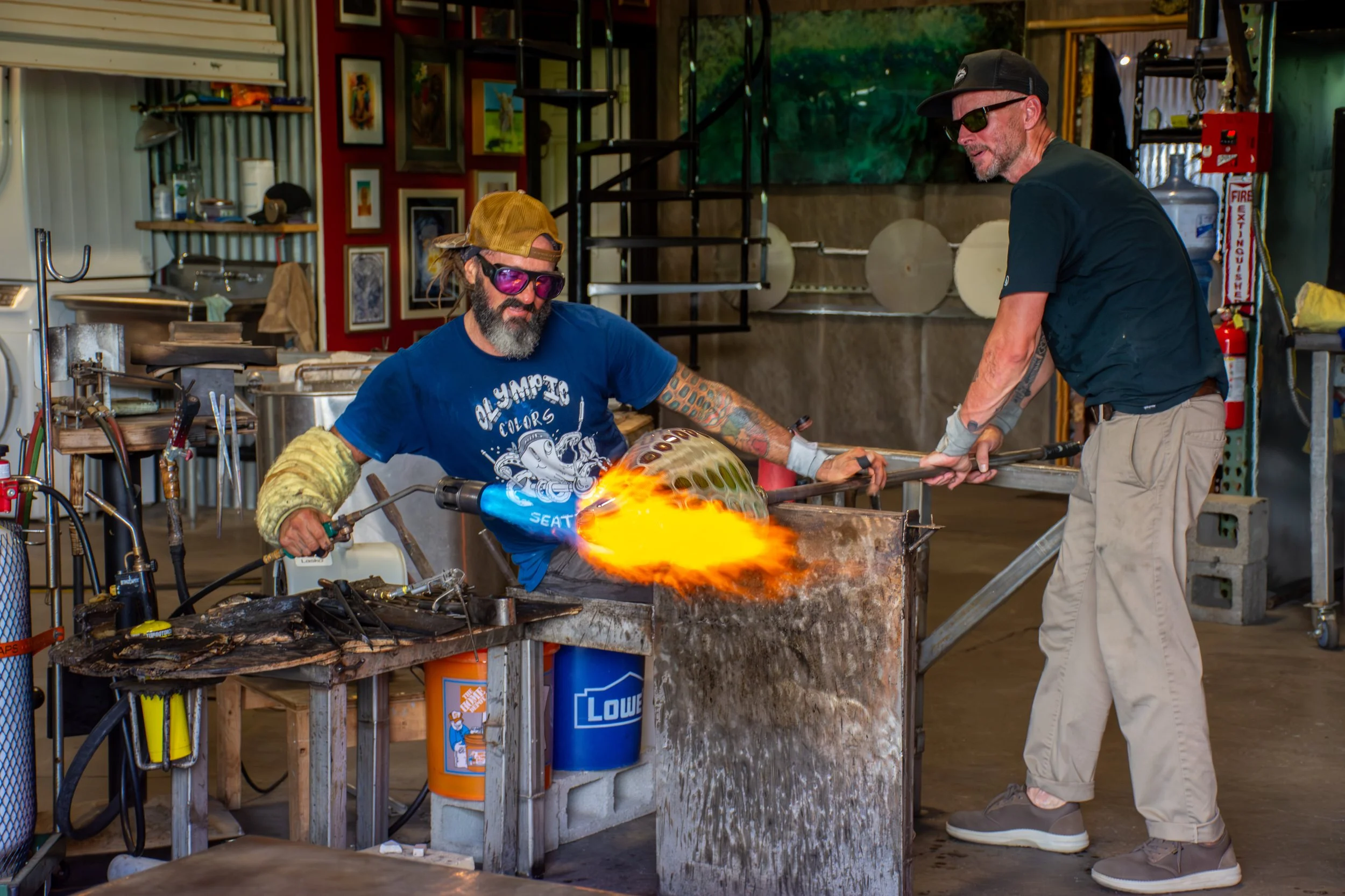 Two men working on glassblowing in a studio, with one man using a torch to shape molten glass and the other assisting, surrounded by art and tools.