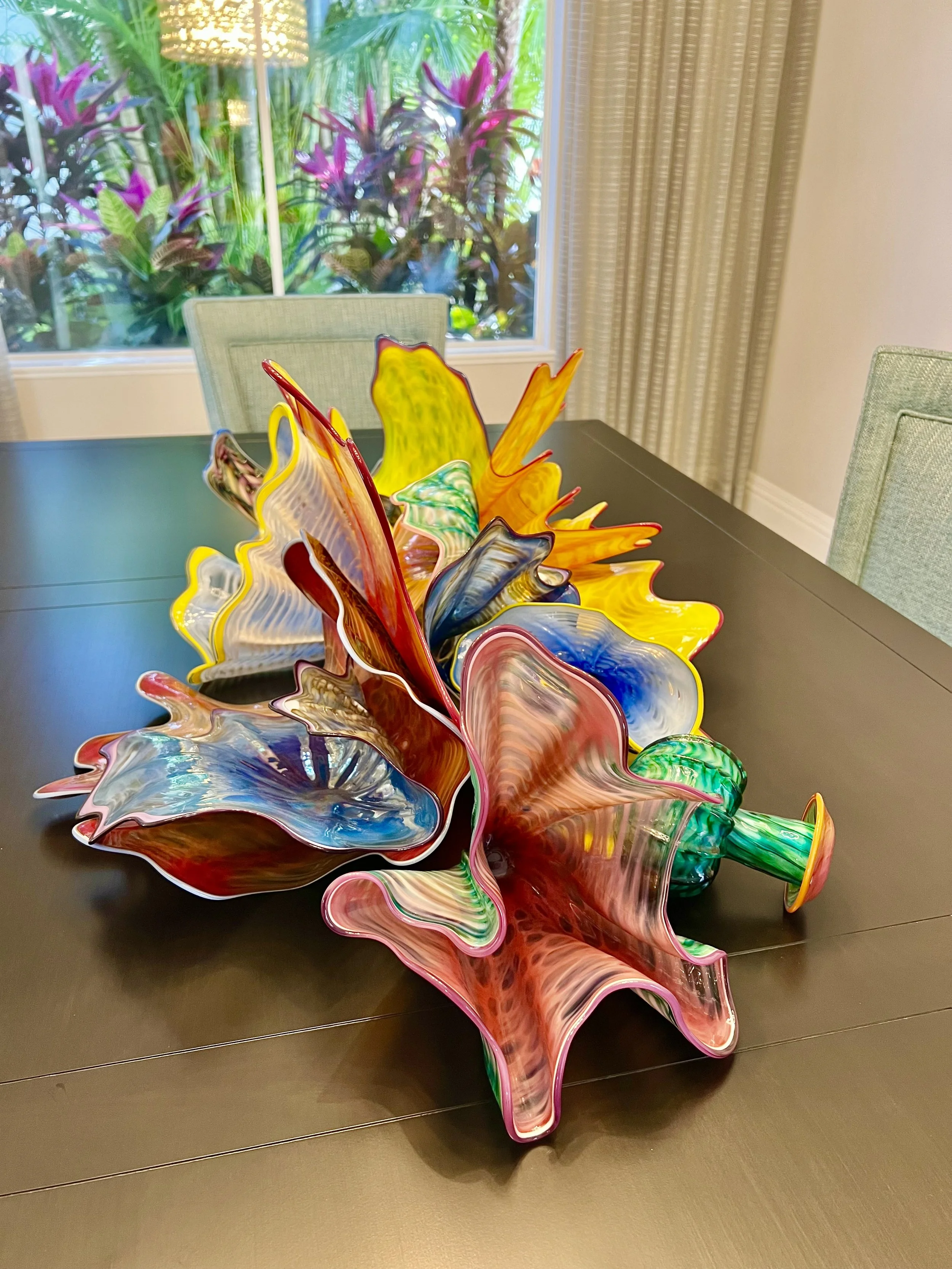 Colorful glass art sculpture on a dark table with a window showing plants and sunlight in the background.