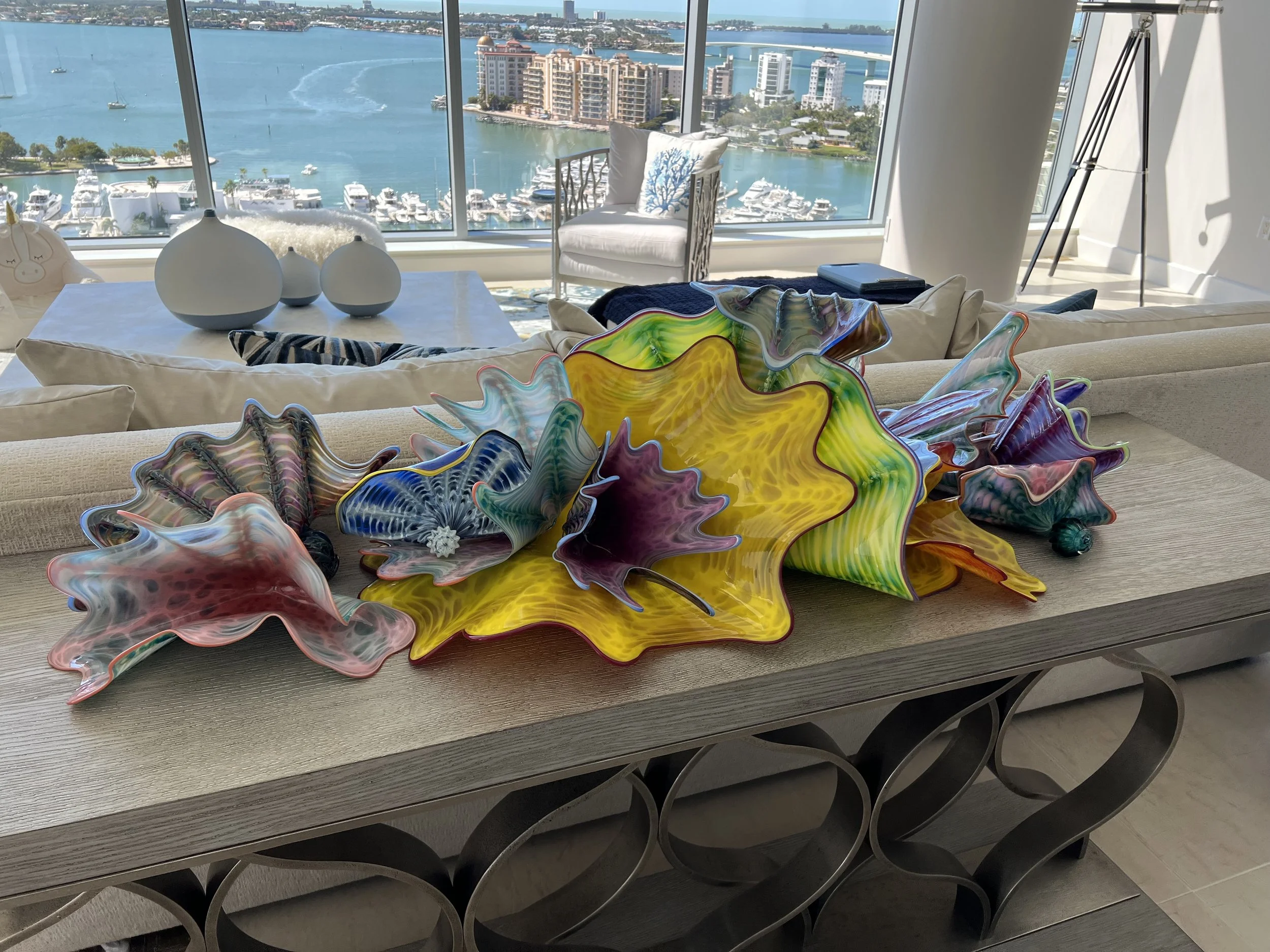 Colorful, decorative glass sculptures resembling large leaves or flowers on a wooden table in a modern apartment with a city and water view through large windows.