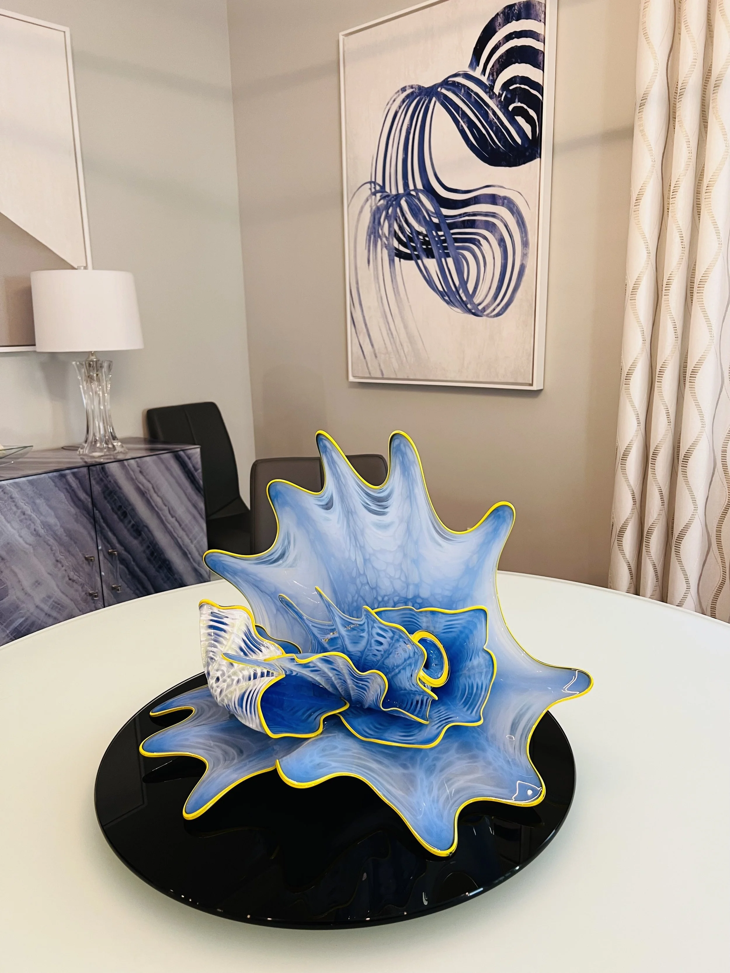 A blue and white abstract glass sculpture with yellow edges, set on a black tray on a white table in a modern room with abstract artwork on the wall and beige patterned curtains.