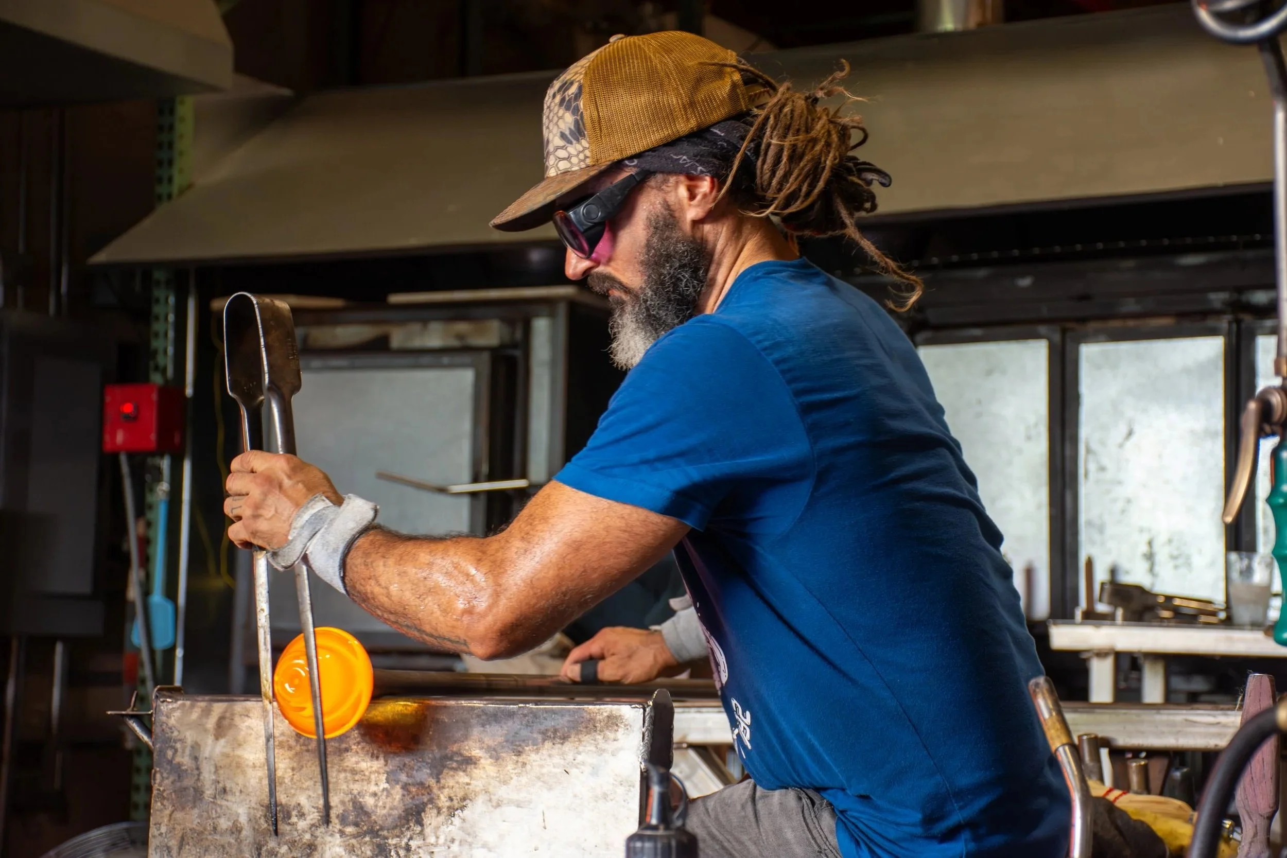 A man with dreadlocks, wearing a baseball cap, sunglasses, a blue t-shirt, and a gray wristband, is grilling over an open flame, using a pair of tongs in a rustic outdoor kitchen.