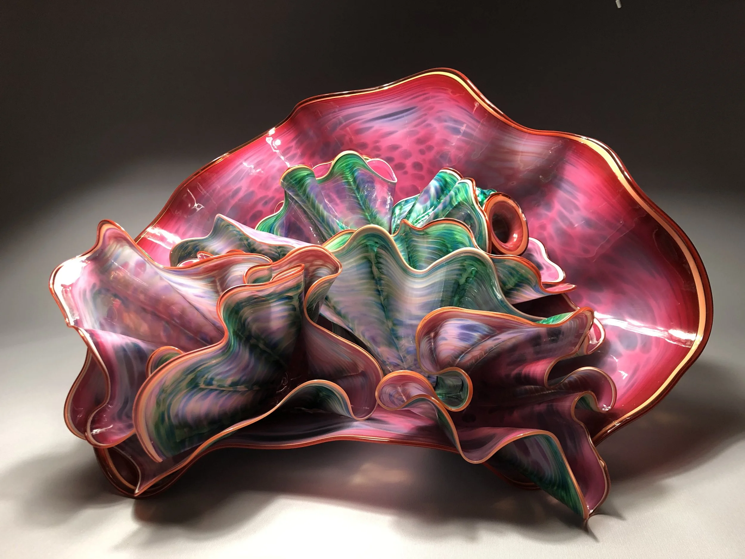 Colorful glass art piece with ruffled, layered petals in shades of pink, green, and purple, edged with gold.