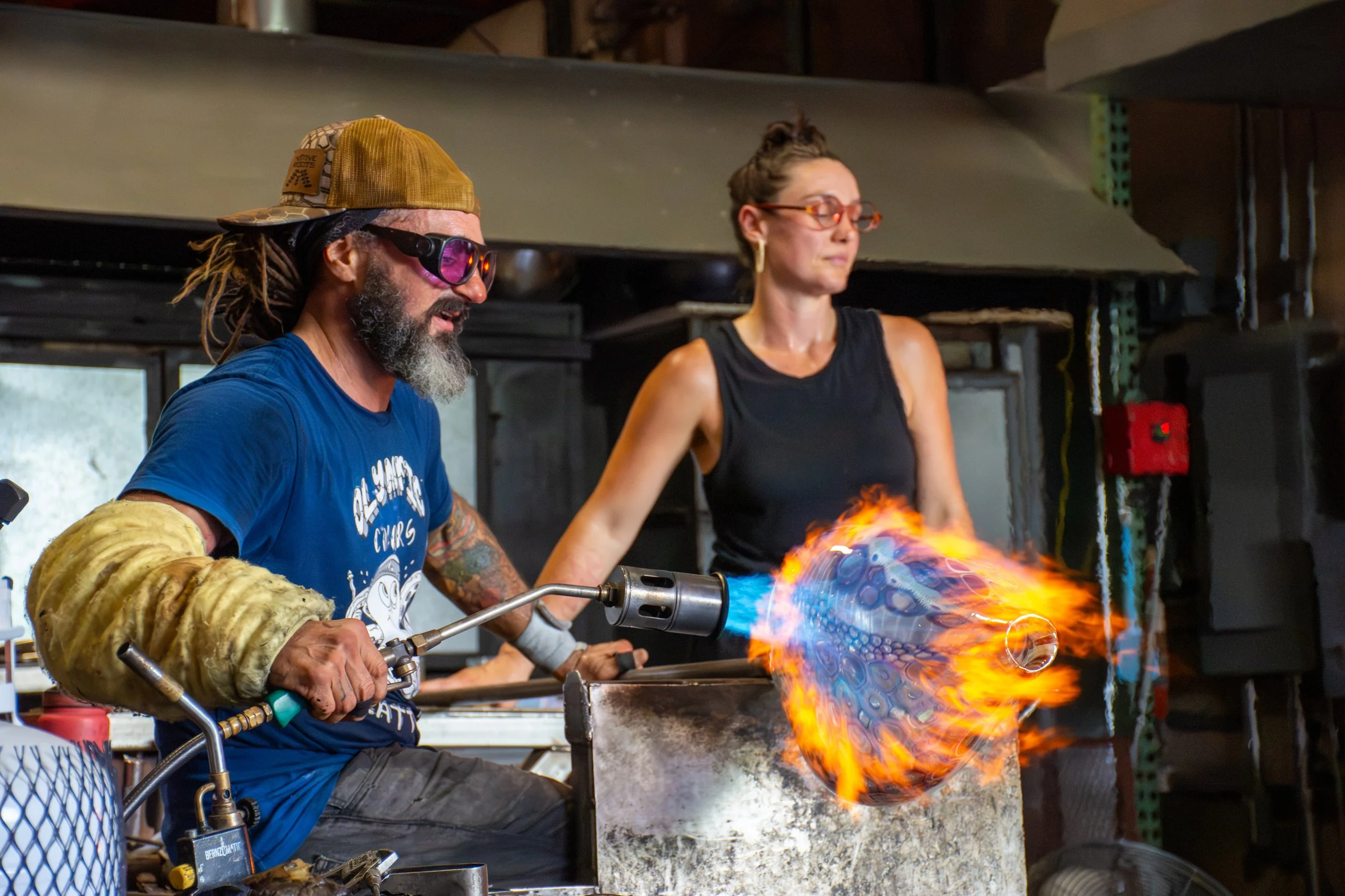 A man with dreadlocks, sunglasses, and a beard uses a blowtorch on a glass piece, creating a flame and colorful glow, while a woman with glasses and a black tank top watches nearby in a workshop.