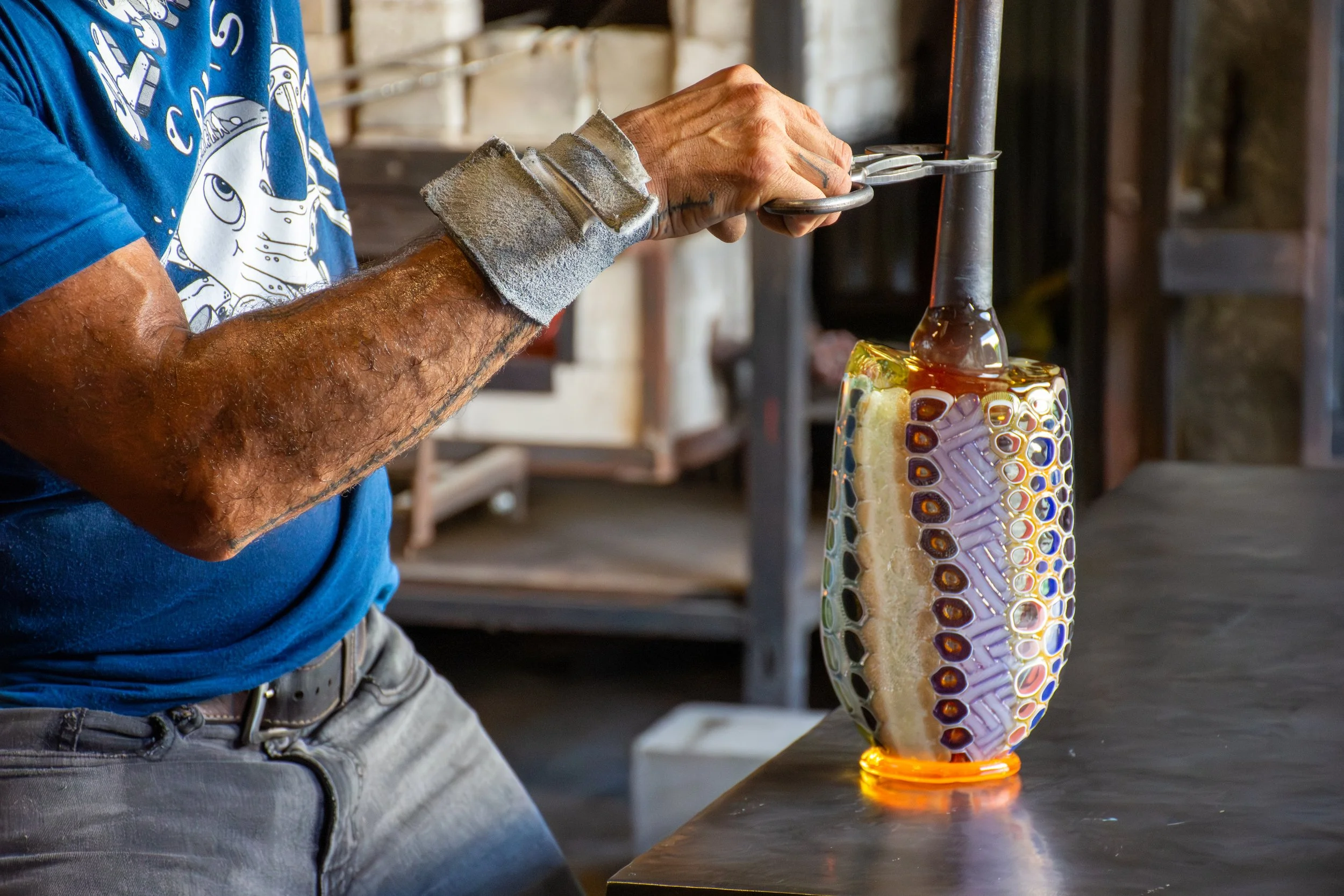 A person shaping colorful glass into a vase using specialized tools in a glass studio.