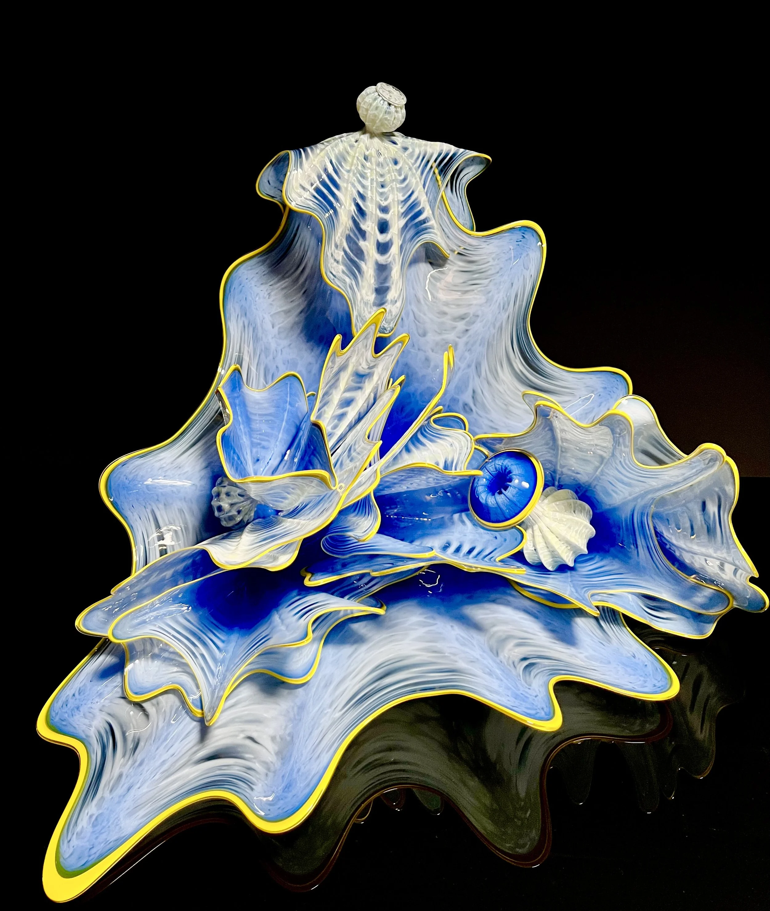A glass art sculpture with blue, white, and yellow accents, resembling a blooming flower, displayed against a black background.