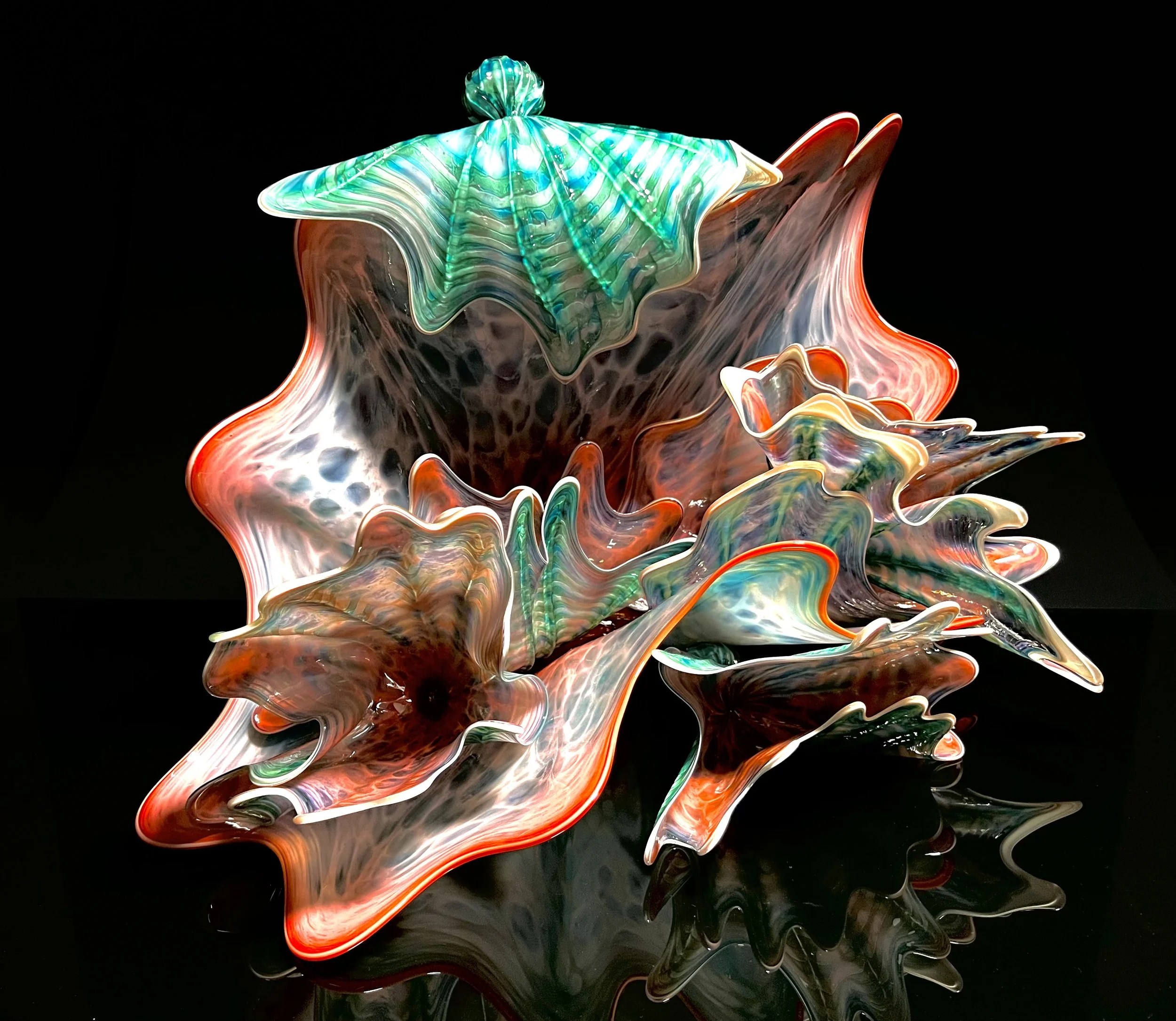 Colorful, abstract glass sculpture with flowing, organic shapes and a black background, reflecting on a glossy surface.