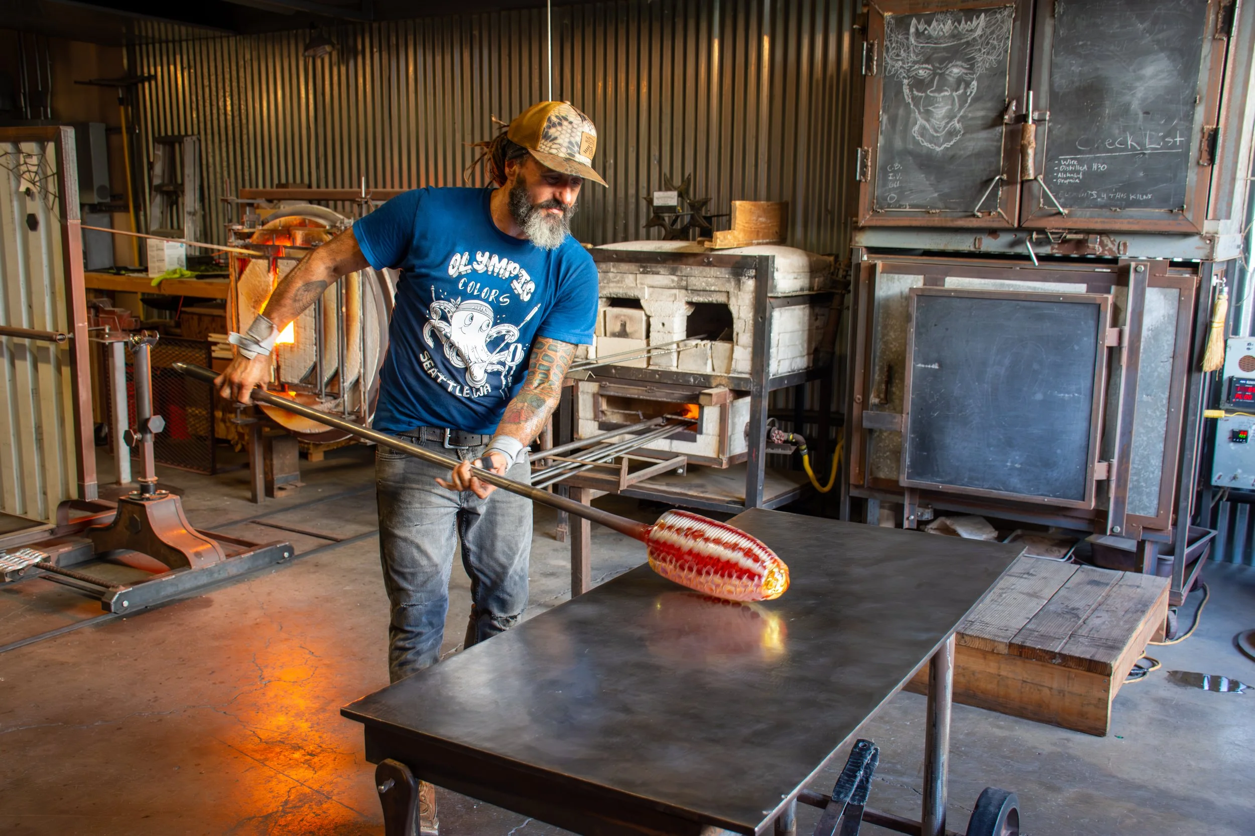 A glassblower is shaping molten glass using a long metal rod in a workshop with industrial equipment and a chalkboard in the background.
