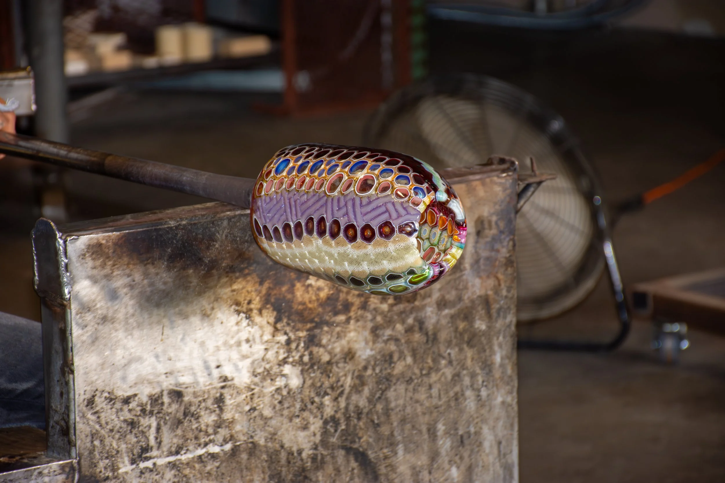 Colorful glass-blown sculpture in process, resting on a metal workbench, with a glassblowing tool attached, in a glass studio.