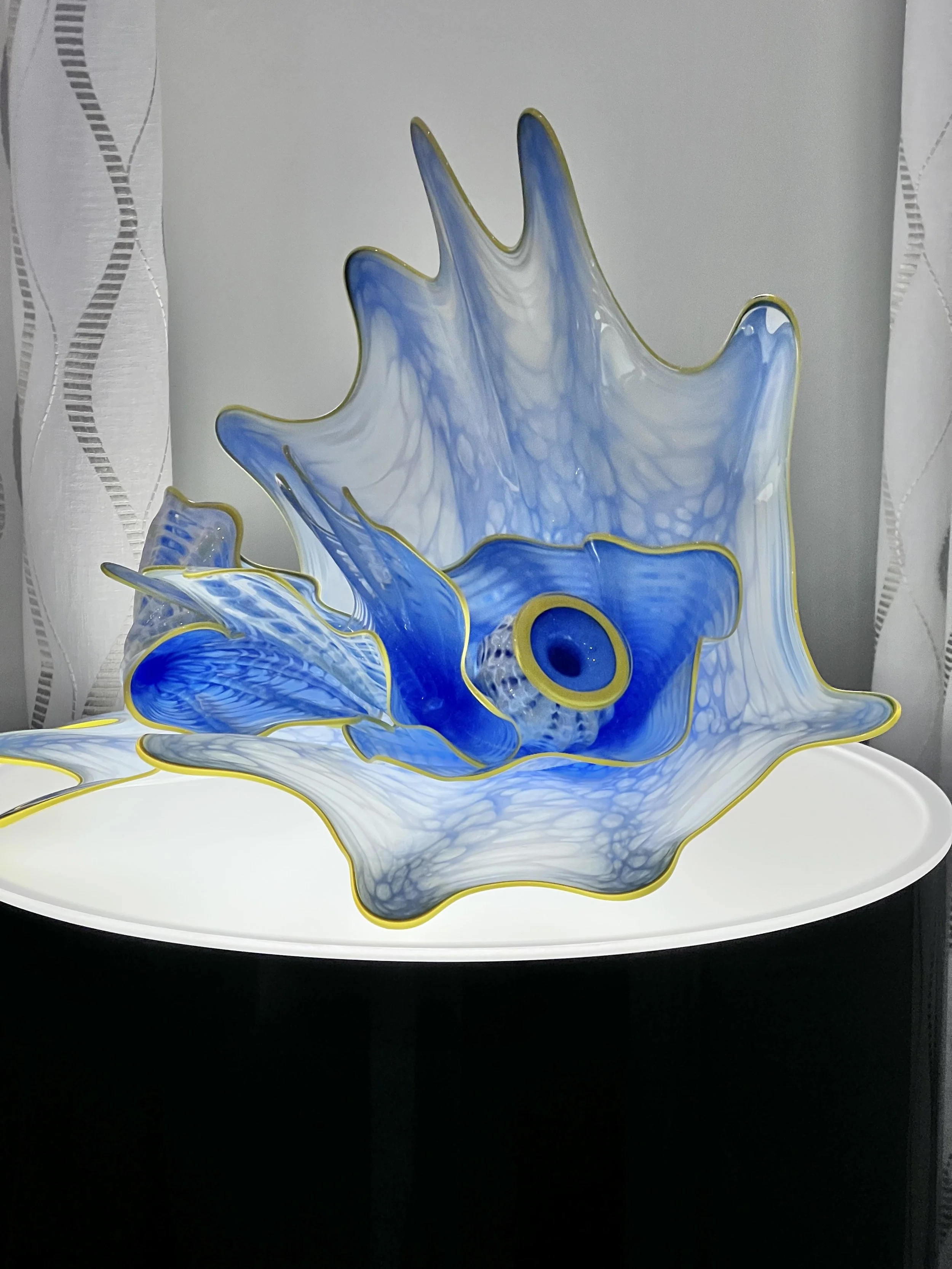 Blue and white glass sculpture with gold accents resembling a sea creature, displayed on a white surface against a gray wall.