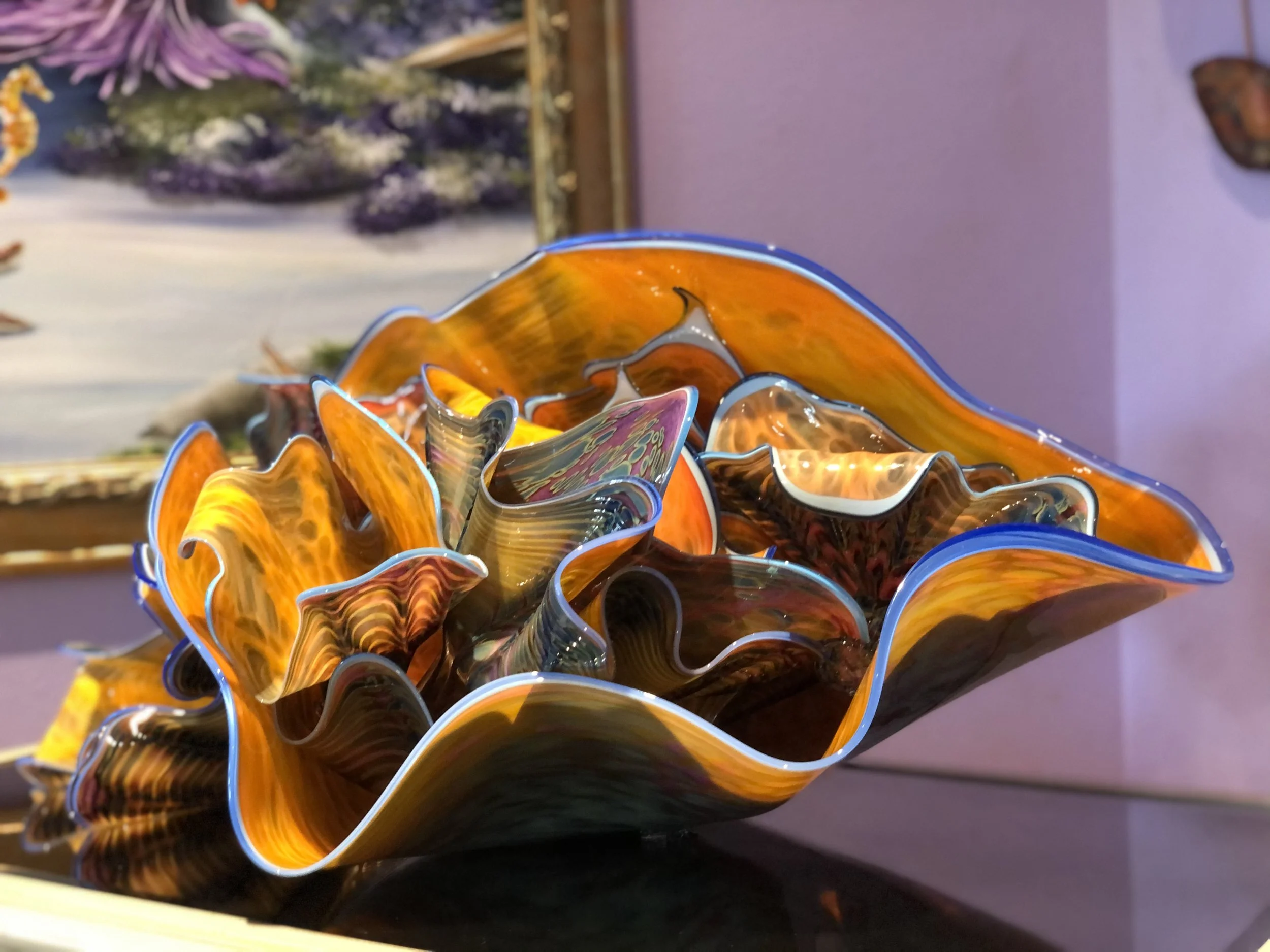 Colorful glass art sculpture with ruffled edges and swirls of orange, yellow, black, and blue, displayed indoors with paintings and purple walls in the background.