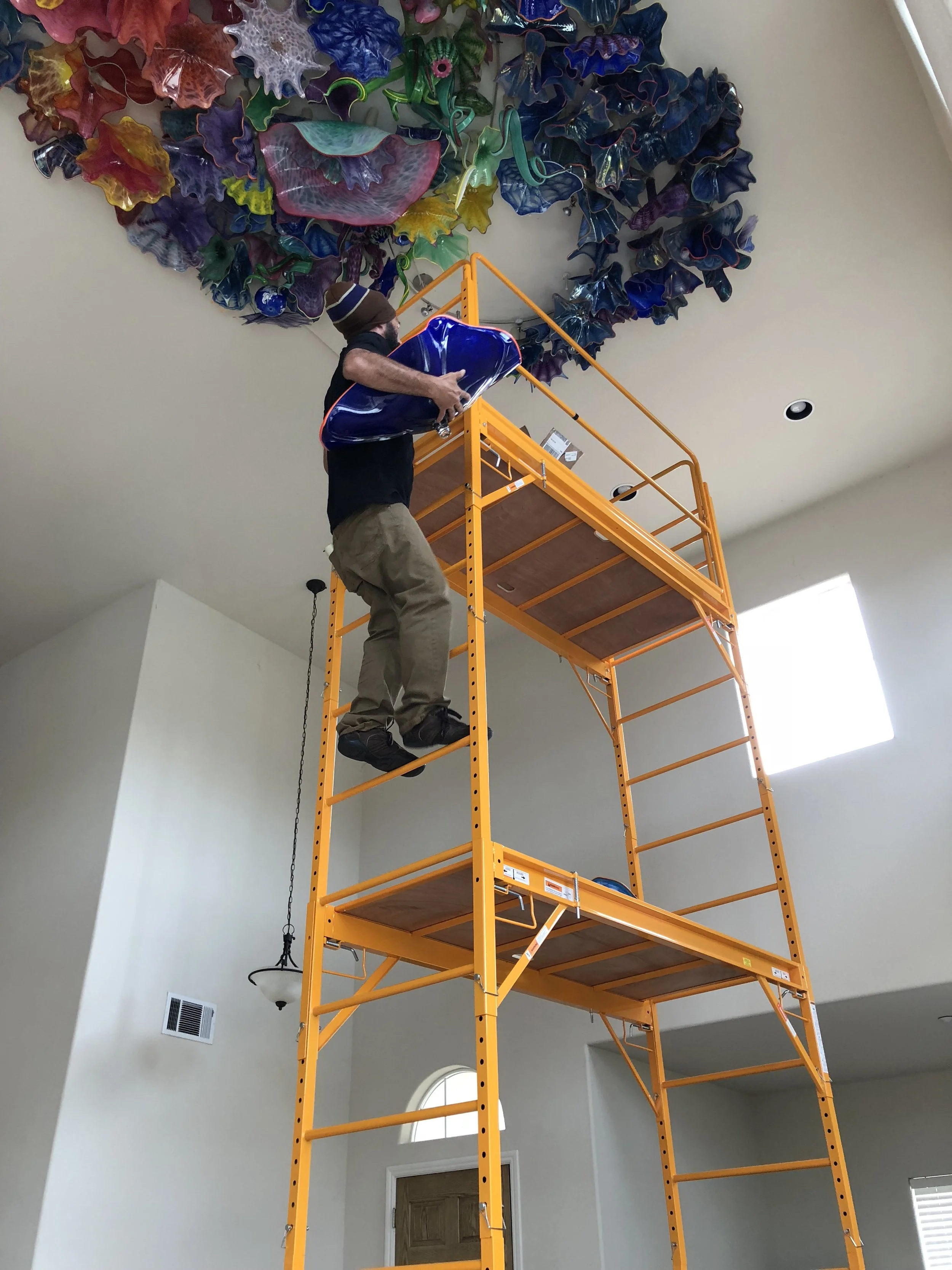 A man standing on an orange scaffolding, working on a large colorful glass art installation on the ceiling of a room.