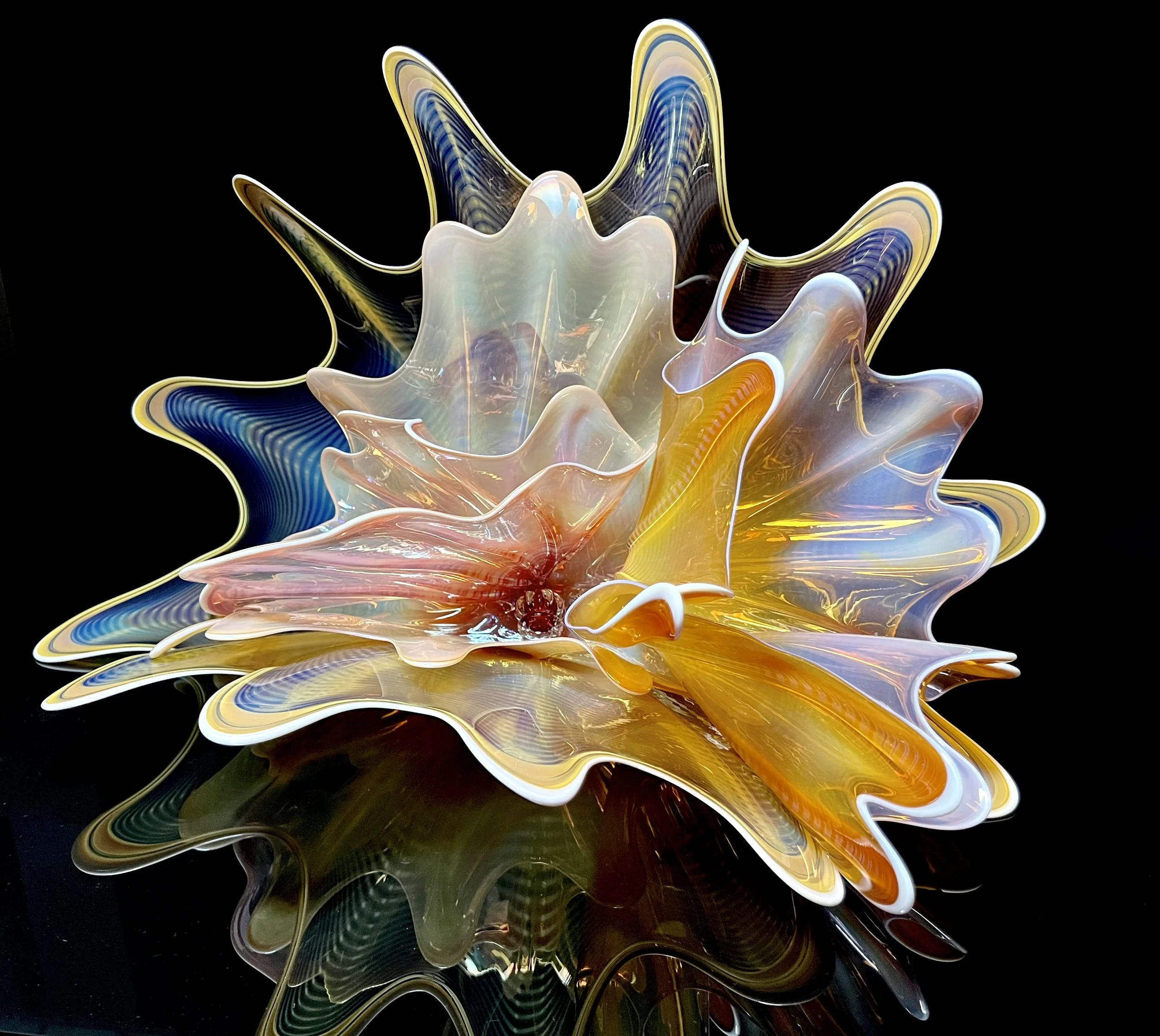 Colorful glass art sculpture with yellow, orange, pink, white, and blue hues on black reflective surface.