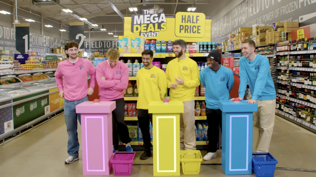 Six people participate in a game show in a supermarket aisle, standing behind three colorful podiums in pink, yellow, and blue, each with a big red buzzer and a basket underneath. The background features signs that say 'Mega Deals' and 'Half Price.'