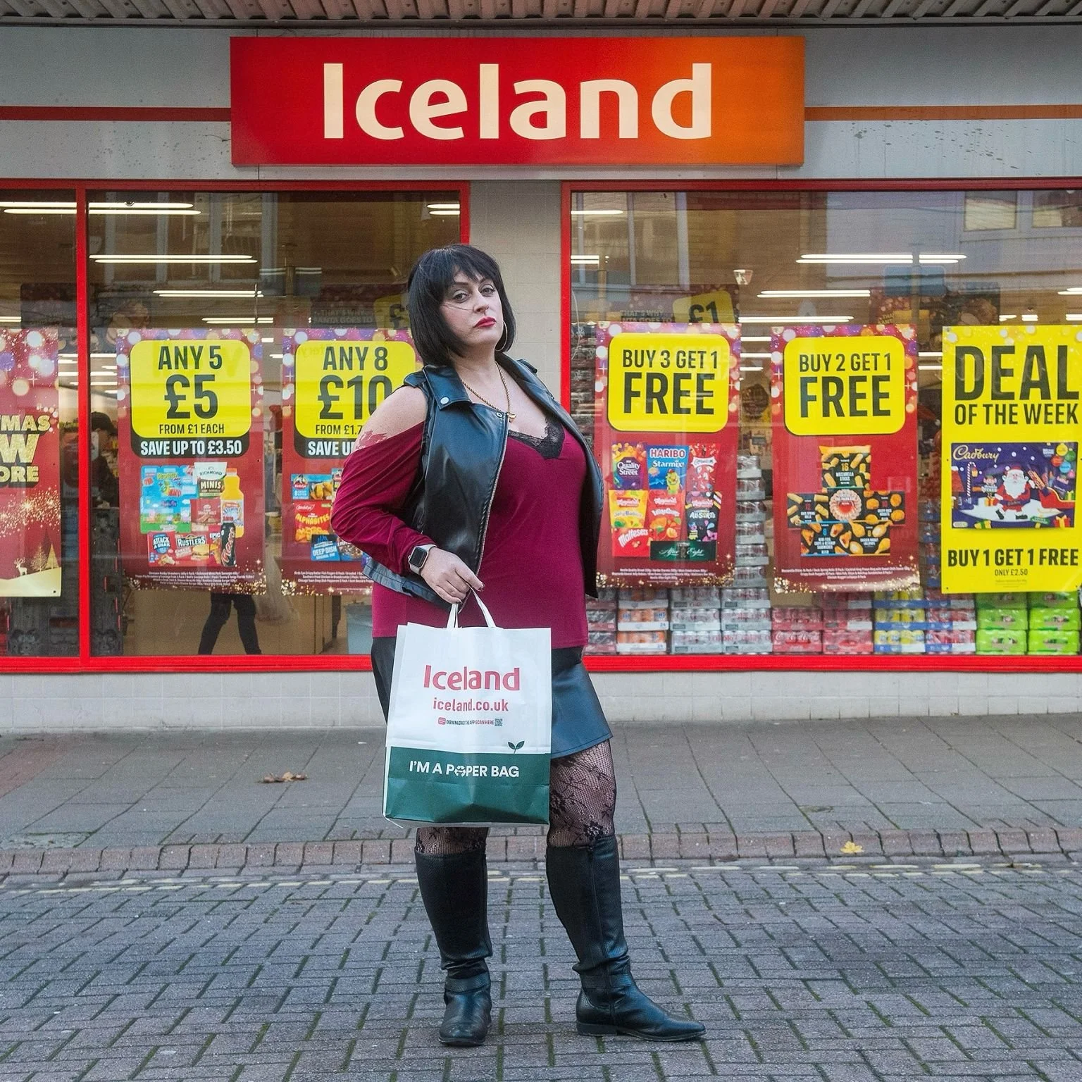 A woman with dark hair and makeup standing outside a store with a large orange and red sign that says 'Iceland.' She is holding a shopping bag that reads 'Iceland' and 'I'm a paper bag.' She is wearing a burgundy dress, a black leather vest, patterned tights, and black boots, with her left hand on her hip.