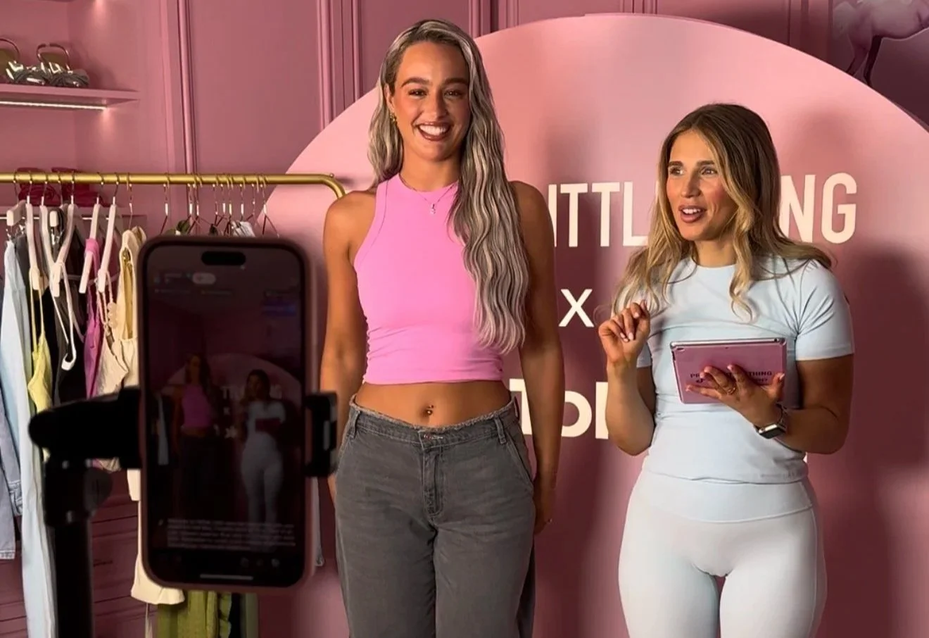 Two women standing in front of a pink backdrop with clothing racks behind them, one woman in a pink top and gray jeans, the other in a white outfit holding a tablet, taking a photo of the first woman.