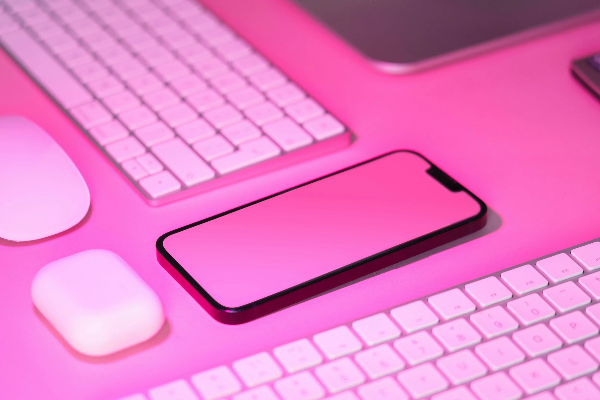 A pink-themed workspace with a wireless keyboard, a smartphone, and a pair of wireless earbuds, all arranged on a pink surface.