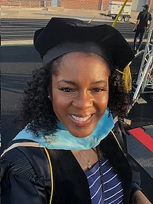 Woman wearing graduation cap and gown, smiling, at outdoor graduation event.