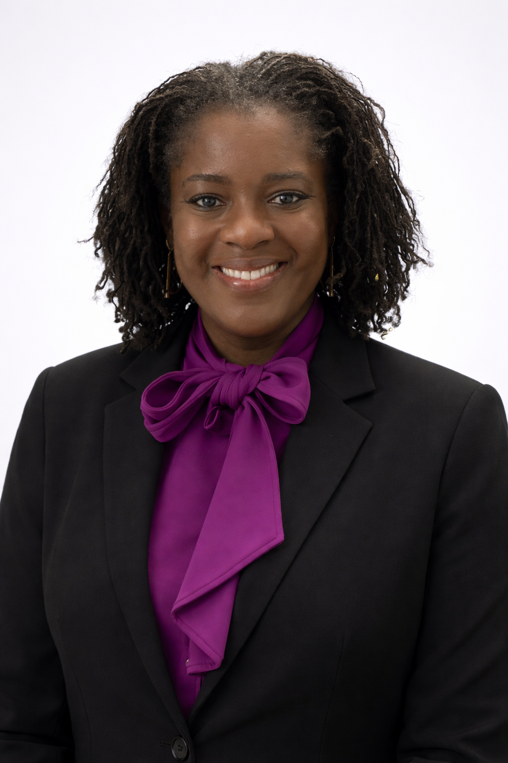 A woman with short curly black hair, wearing a black blazer and a purple blouse with a matching purple bow tie, smiling against a plain white background.