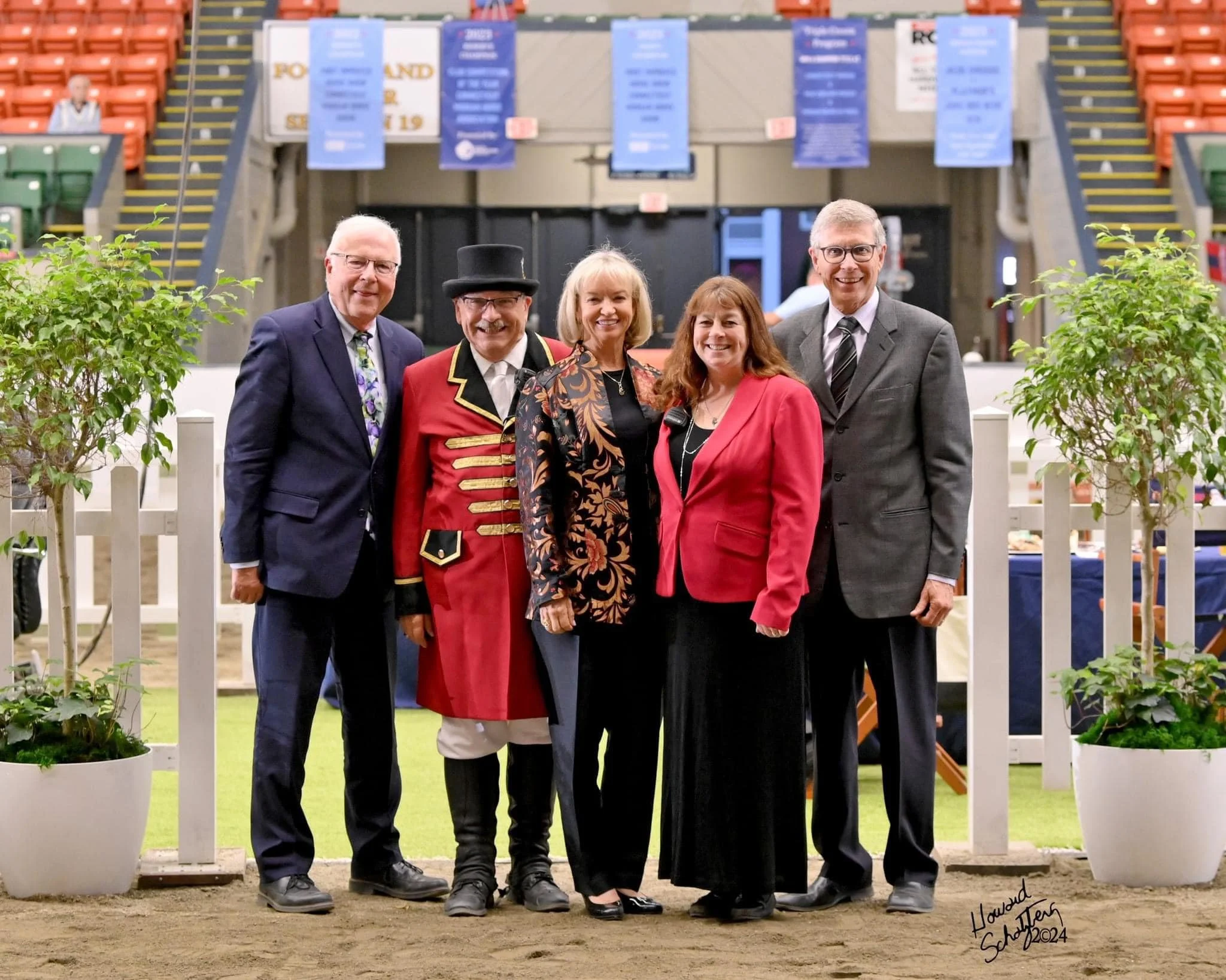 Group of five people standing together at an indoor event, with a circus or fair-like background featuring banners and orange tiered seating, surrounded by potted plants.