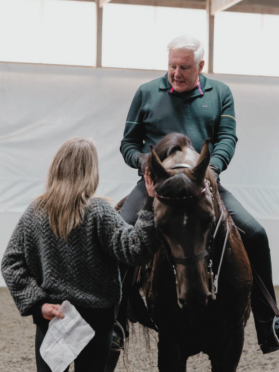 An elderly man with white hair riding a horse inside an indoor arena, while a woman with long blonde hair in a gray sweater stands beside the horse, holding its head.