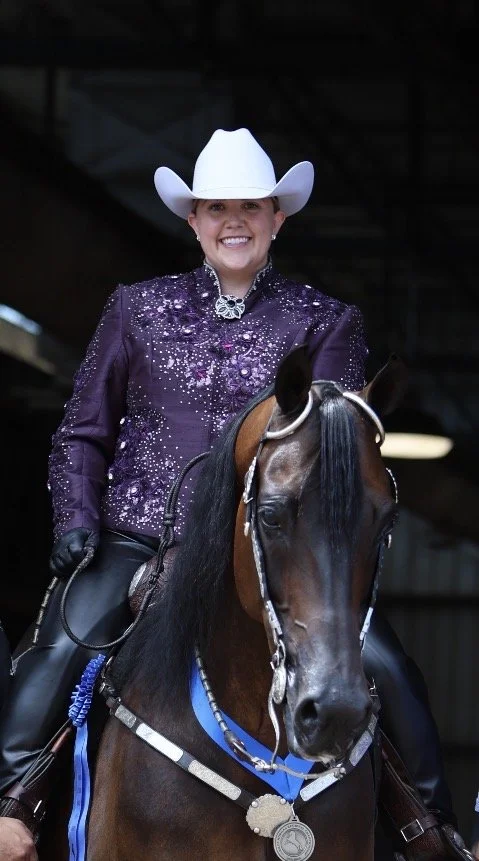 Woman in purple sparkly western attire with white cowboy hat riding a horse with a silver medal around its neck.