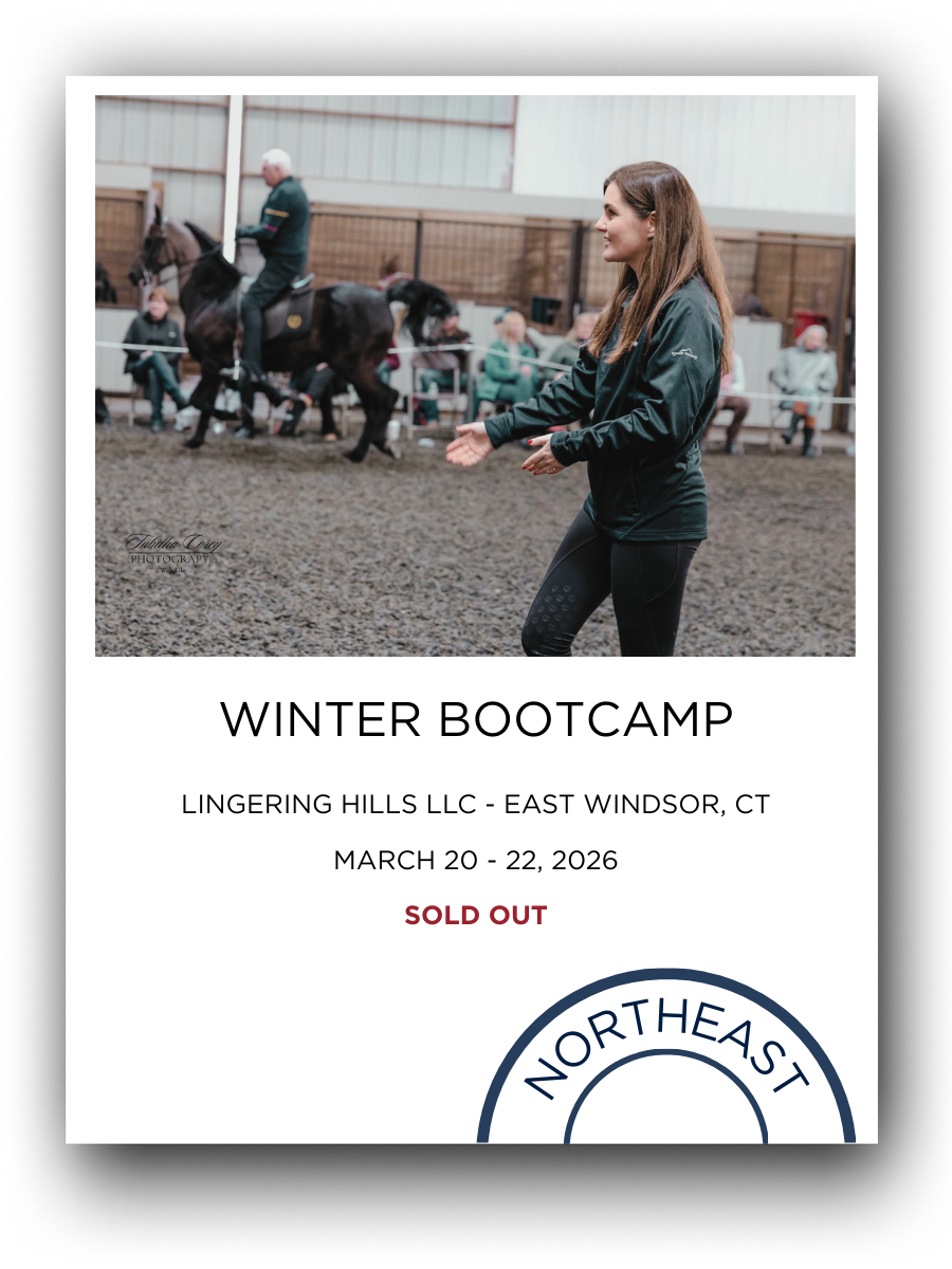A woman standing and smiling during winter bootcamp at an indoor horse riding arena, with a man riding a horse in the background and spectators seated behind a fence.