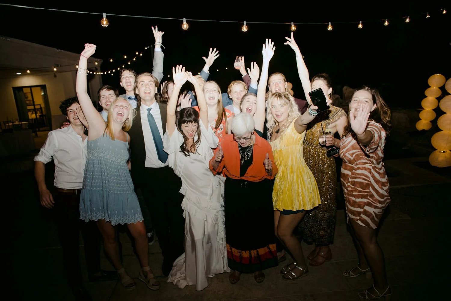 Group of people celebrating at night with string lights, raising their hands, smiling, and taking photos.