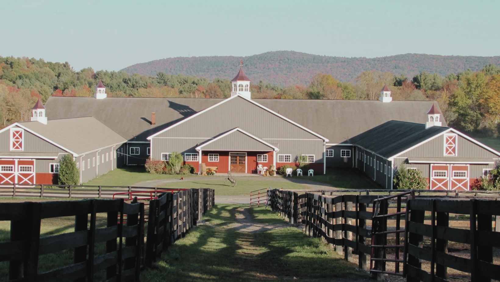 A large barn with multiple sections, gray and red exterior, and decorative white trim, surrounded by green grass, with a scenic background of trees and hills.