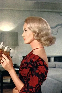 A woman with blonde, vintage-style hair holding a glass of drink, dressed in a red and black floral dress, in a living room setting.