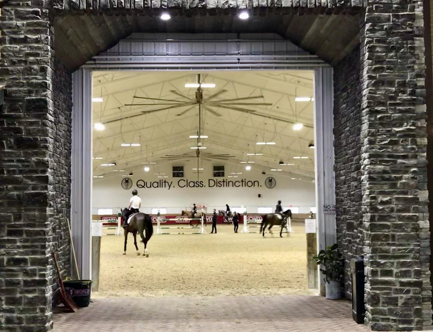 View of an indoor horse riding arena through an opening with brick pillars and a stone arch. Riders on horseback and a few people standing in the arena. Interior has high ceiling with fans, lights, and slogan on wall reading 'Quality. Class. Distinct
