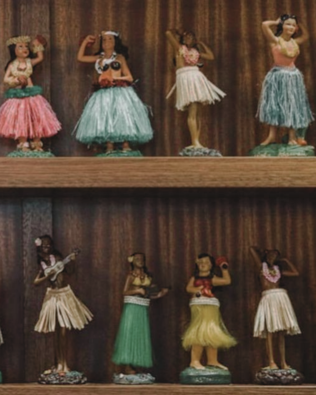 A collection of Hawaiian hula girl figurines displayed on wooden shelves, dressed in colorful grass skirts, leis, and accessories, representing traditional Hawaiian hula dancers.