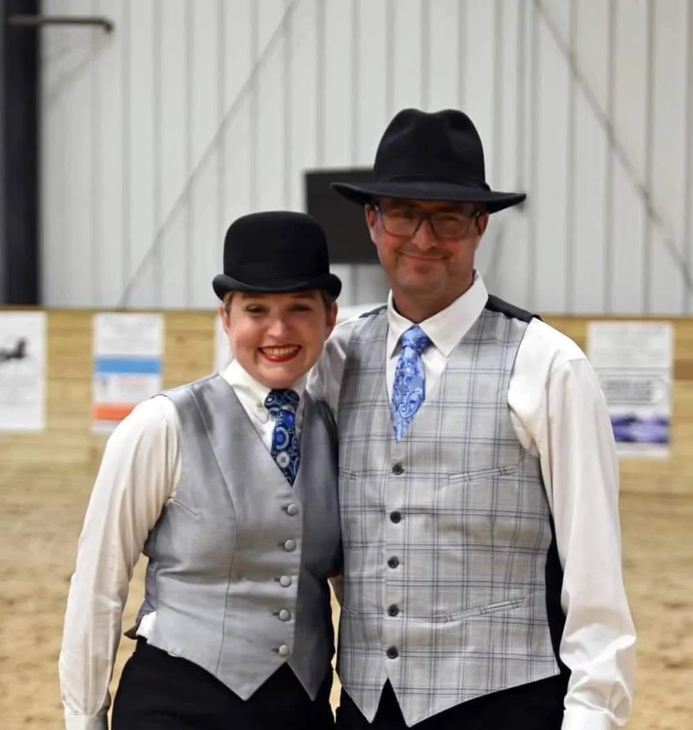 Two people dressed in vintage attire, wearing black bowler hats, vests, white shirts, and patterned ties, standing inside a building with a rustic background.