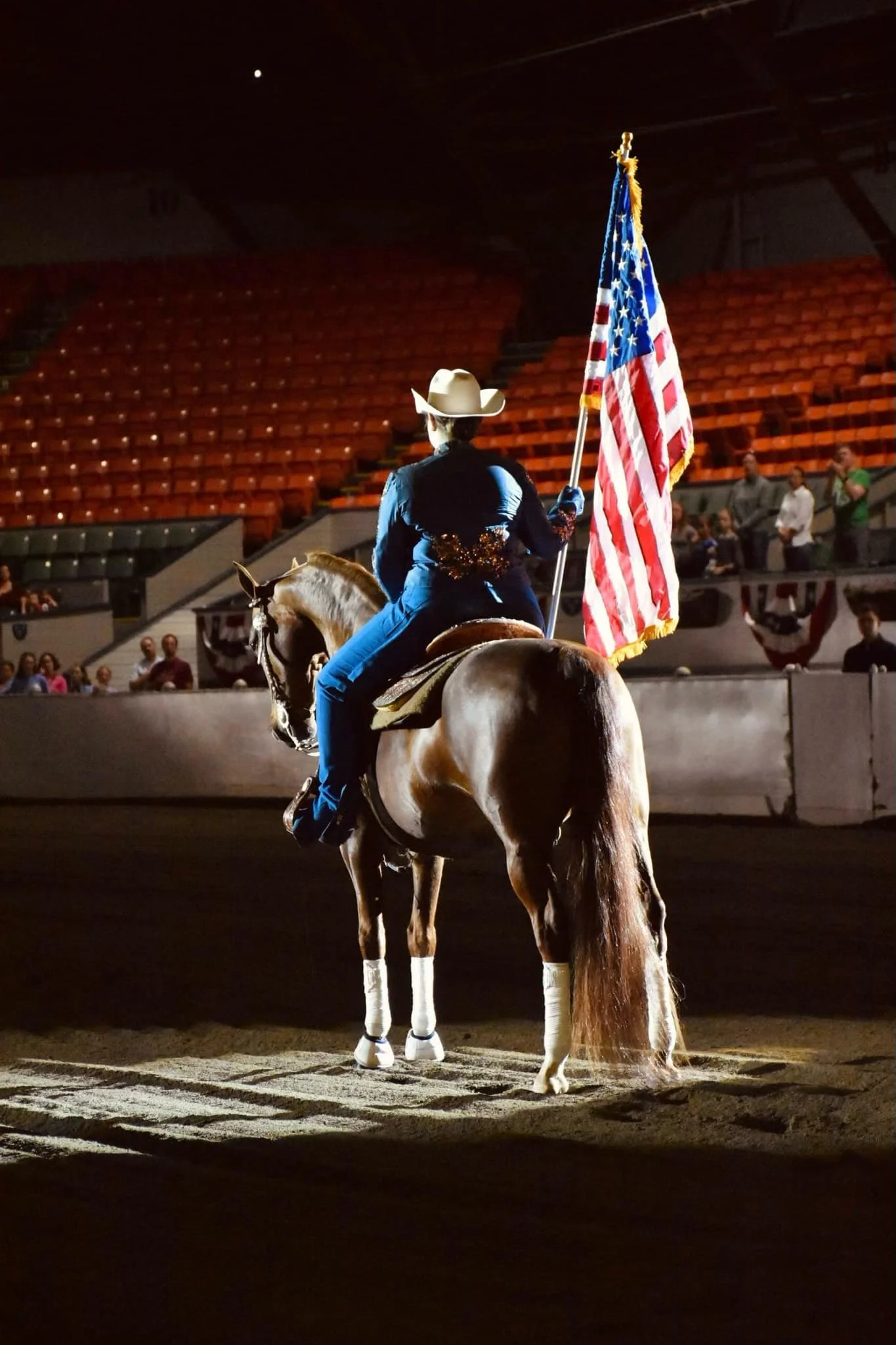 A person dressed as a cowboy riding a horse in an indoor arena, holding an American flag, with empty red seats and a few spectators in the background.
