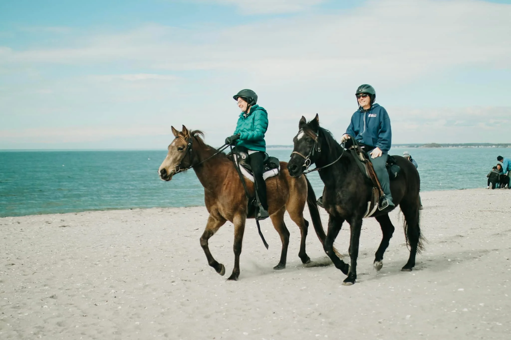 Two women riding horses along a beach with ocean in the background, wearing helmets and jackets.
