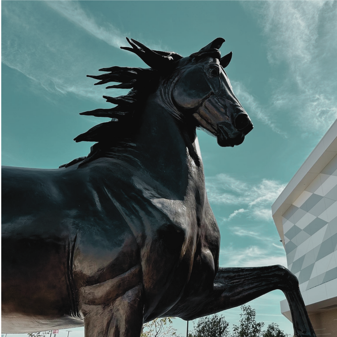 Large black horse statue with flowing mane set against a sky with clouds and a modern building in the background.